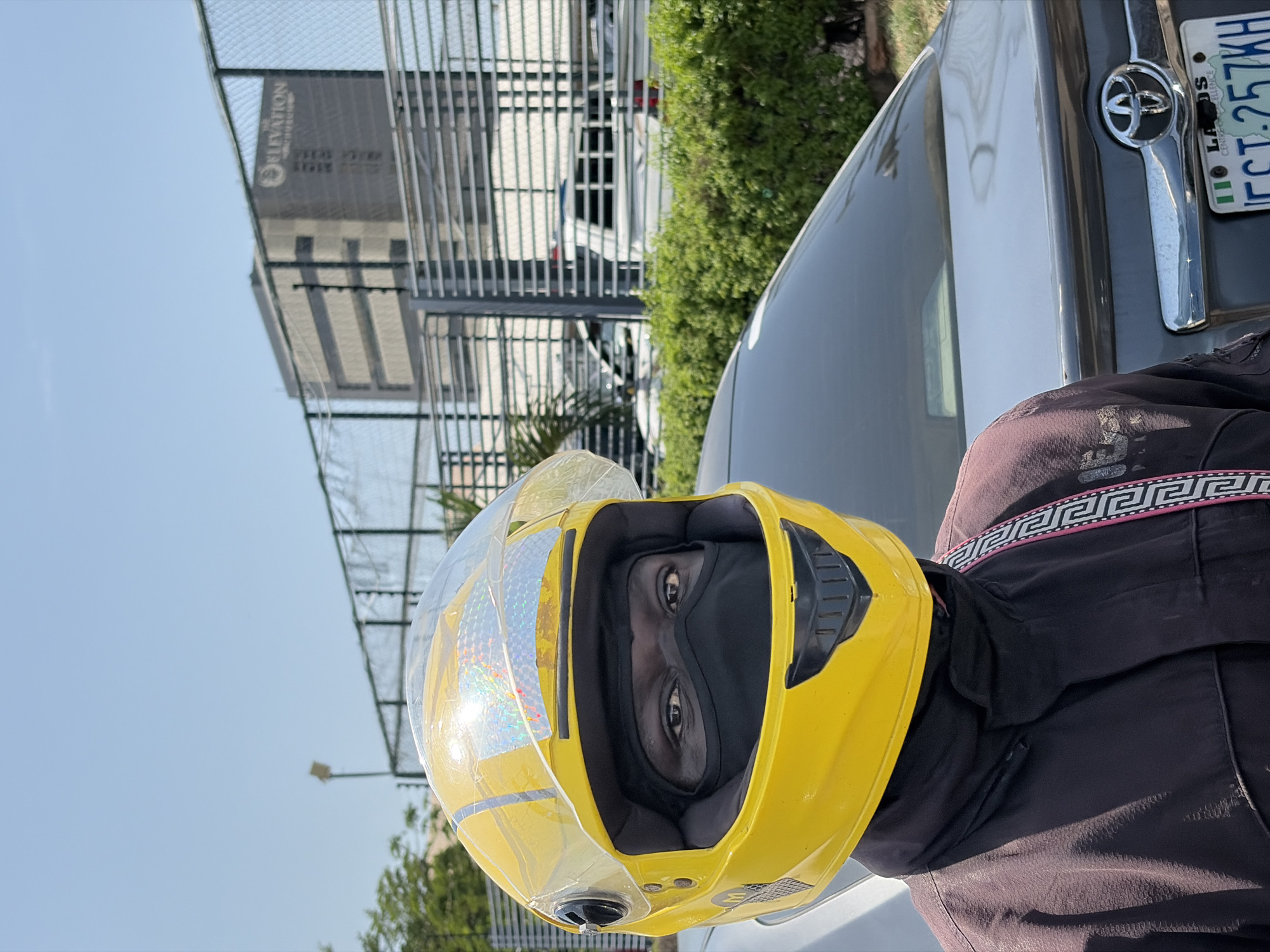 A close-up, rotated shot captures a person wearing a yellow motorcycle helmet with a clear visor, positioned next to a gray Toyota car, its license plate displaying "LAGOS FCT.257KH". The person wears a dark garment with a patterned strap. In the background, there are partially visible trees and a multistoried building with "ELEVATION" signage, implying an urban setting, identified as Igboefon, Nigeria.