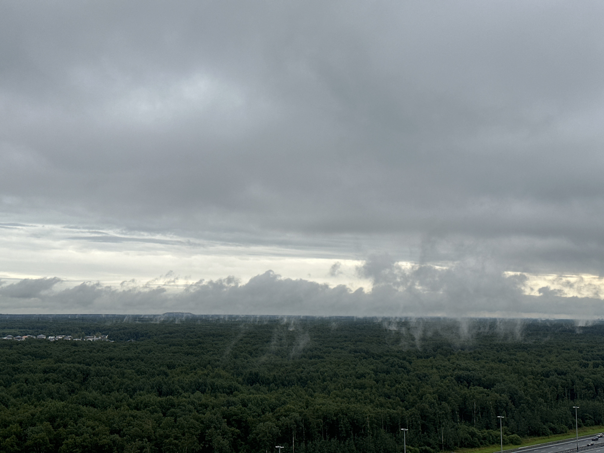 An aerial, wide shot captures a vast, dense forest under a heavily overcast sky. The trees are predominantly dark green, with some lighter green foliage interspersed. In the distance, a cluster of houses with white walls and darker roofs is visible, nestled within the treeline. Above the horizon, a band of low-lying clouds or mist hangs over the forest, creating a hazy effect. Streaks of light pierce through breaks in the clouds in the upper portion of the frame. To the right of the image, a portion of a multi-lane road is visible, with a few cars driving on it, and streetlights line the side of the road. The scene is set in the city of P.im.sverdlova, Russia.