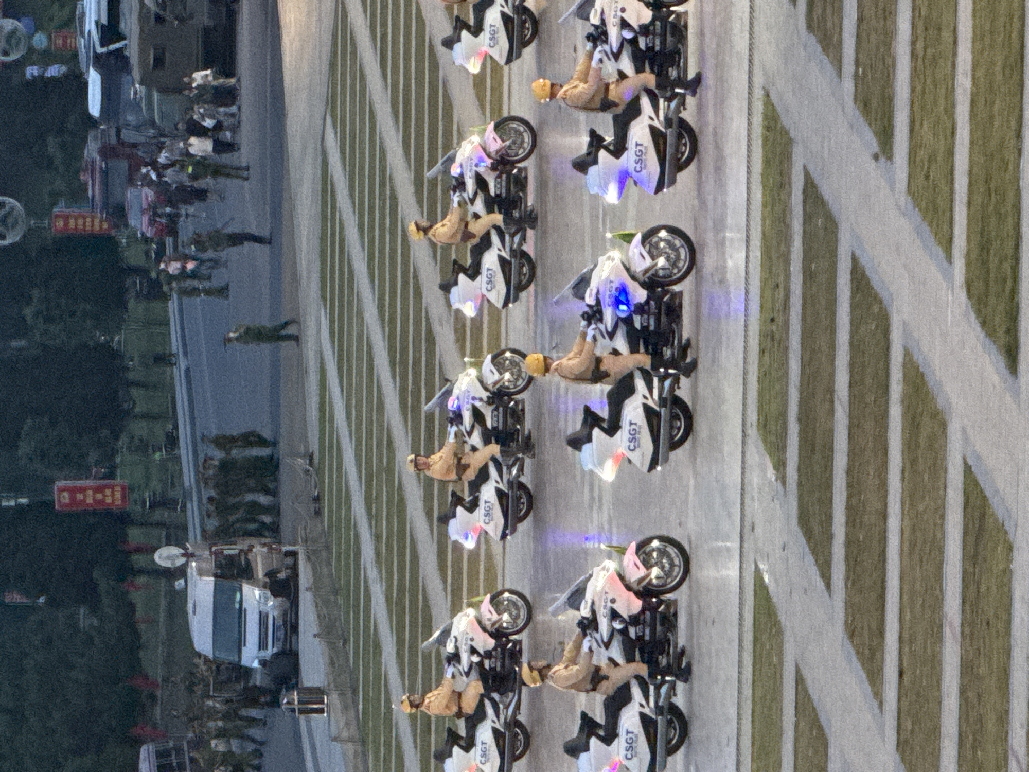 The image depicts a wide-angle view of a street or public square in Da Lat, Vietnam, during daylight hours. The foreground features a patterned ground composed of alternating strips of grass and concrete walkways. In the midground, several police officers on motorcycles are positioned in rows. These motorcycles are white with pink accents and have "CSGT" visible on the sides, indicating Traffic Police. The officers are wearing light brown uniforms and yellow helmets. In the background, a white van with its doors open is parked on the street, surrounded by a group of people, some in uniform and others in civilian clothing. Further in the background, trees and buildings are visible, along with other vehicles including a larger transport vehicle. The scene appears to be an organized event or a public gathering, possibly a parade or a security detail.