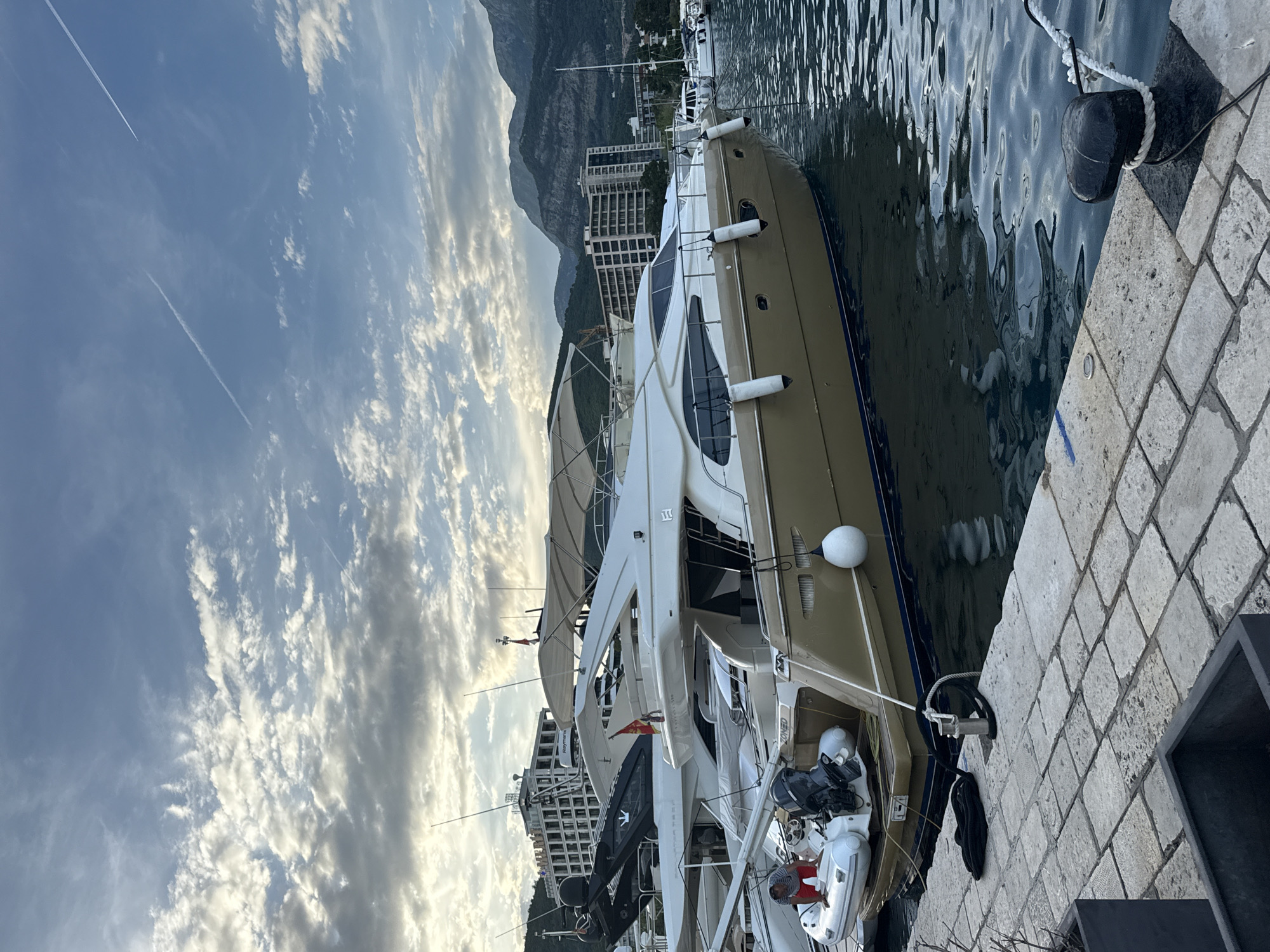 The image depicts a marina scene in Budva, Montenegro, under a partly cloudy sky, likely during late afternoon or early evening.
In the foreground, a light grey stone-paved pier occupies the bottom section. Visible on the pier are two rectangular dark grey planters (partially cropped), coiled black mooring lines, and a black cylindrical mooring bollard with a white rope wrapped around it. Another lighter grey, cylindrical mooring cleat is further back, also with a rope attached.
The midground is dominated by a luxurious motor yacht with a distinctive gold-tan hull and a white superstructure. Several white cylindrical fenders are suspended along its port side. At the stern of this yacht, a white inflatable tender with a black outboard motor is attached. A man wearing a striped shirt and red shorts is standing in the tender, appearing to be engaged with it. On the yacht's superstructure, two small flags are visible; one is red with a golden double-headed eagle, consistent with the flag of Montenegro. Behind this main yacht, other white luxury yachts are docked, one of which displays a "Raymarine" radar dome on its flybridge and a stylized crown logo on its hull. The water in the marina is dark blue-green, exhibiting gentle ripples and reflections of the boats and sky.
The background features several multi-story, modern buildings, likely hotels or residential complexes, with numerous balconies and windows, situated along the coastline. One building also has a "Raymarine" dome on its roof. Directly behind these structures, large, rugged mountains with varied rocky and forested textures rise prominently, creating a dramatic backdrop. A sailboat mast is faintly visible on the far right, blending with the distant landscape.
The sky above is a light blue with scattered white and grey cumulus and stratocumulus clouds, indicating partial cloud cover. Two distinct white contrails from aircraft are visible in the upper sky, one extending diagonally from the upper right and another fainter one further left. The lighting is soft and diffused, suggesting indirect sunlight or the approach of dusk.