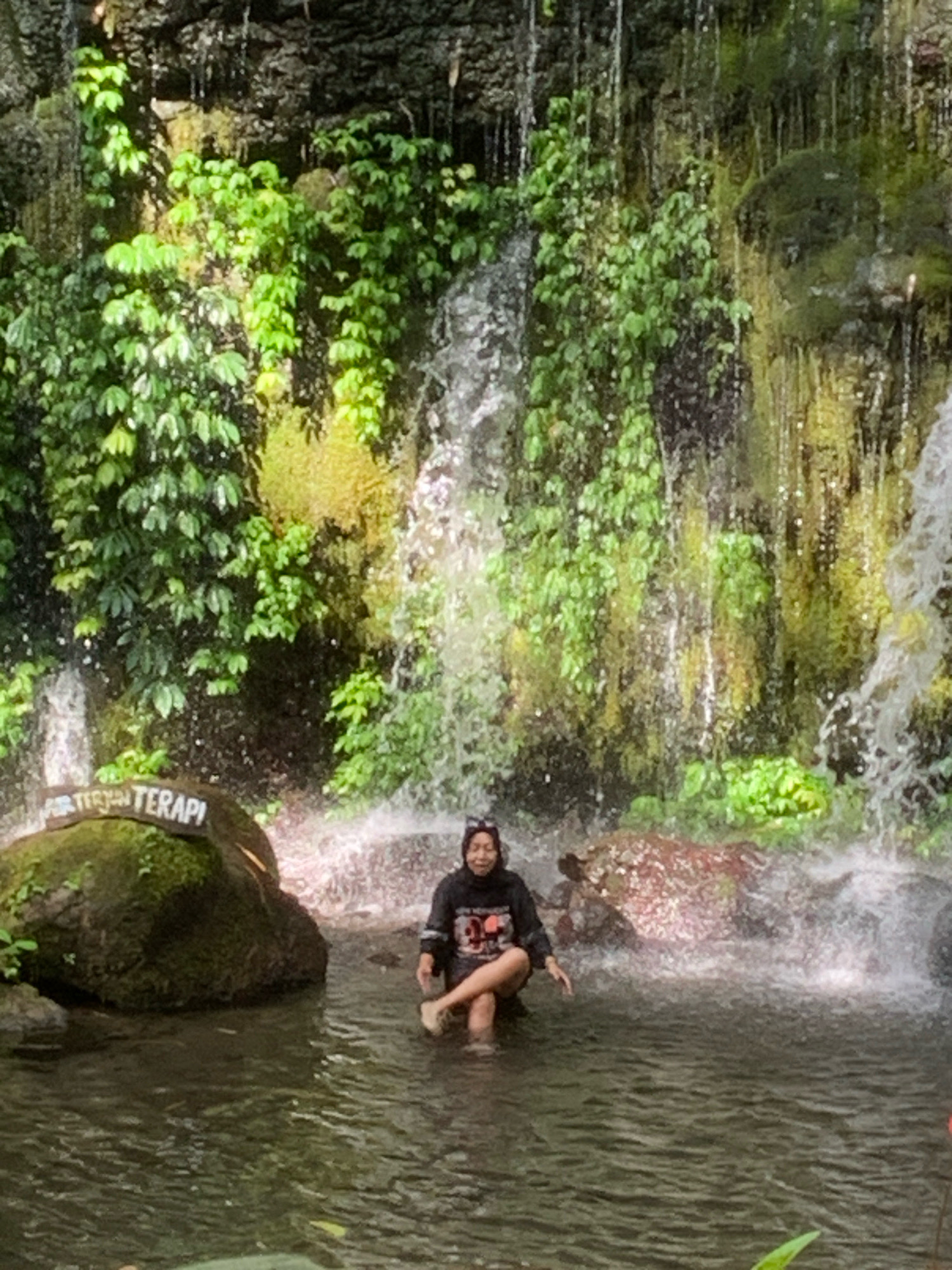 The image depicts a person sitting in a shallow body of water in front of a waterfall in Pronojiwo, Indonesia. The waterfall cascades down a rocky cliff face covered in lush green vegetation. To the left of the frame, a large rock with the inscription "AIR TERJUN TERAPI" (meaning "Therapy Waterfall") is visible. The person, wearing a black headscarf and a black sweatshirt with graphics, is seated with their legs crossed, partially submerged in the water. The water appears clear and reflects the surrounding foliage and the spray from the waterfall. The overall scene suggests a natural, tranquil environment.