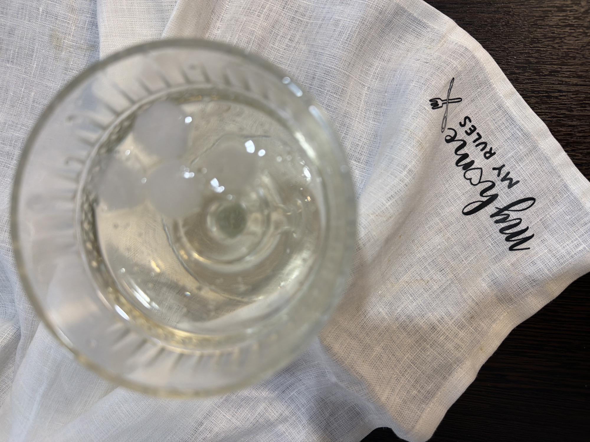 A close-up, overhead shot shows a glass of clear liquid with ice cubes sitting on a white linen cloth. The cloth is draped over a dark wooden surface. The glass has a ridged pattern around its rim. The white cloth has a black graphic printed on it, which includes text that reads "my home MY RULES" and a drawing of a fork and spoon. The image does not contain any elements that would suggest a location in Chaikovsky, Russia.