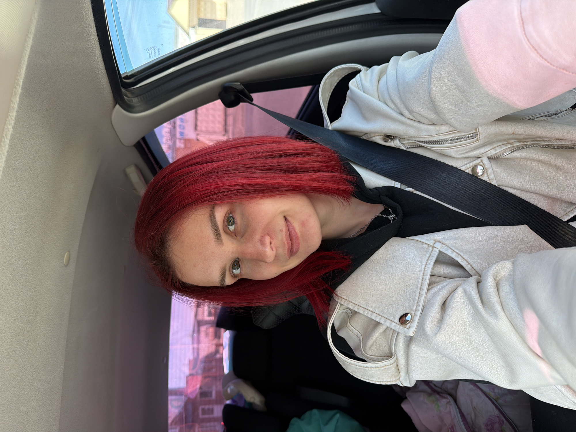 A woman with red hair is wearing a seatbelt and a light-colored leather jacket in what appears to be the interior of a car. A building is visible through the car window; judging by the presence of buildings of that type, it appears to be within the city of Chaikovsky, Russia.