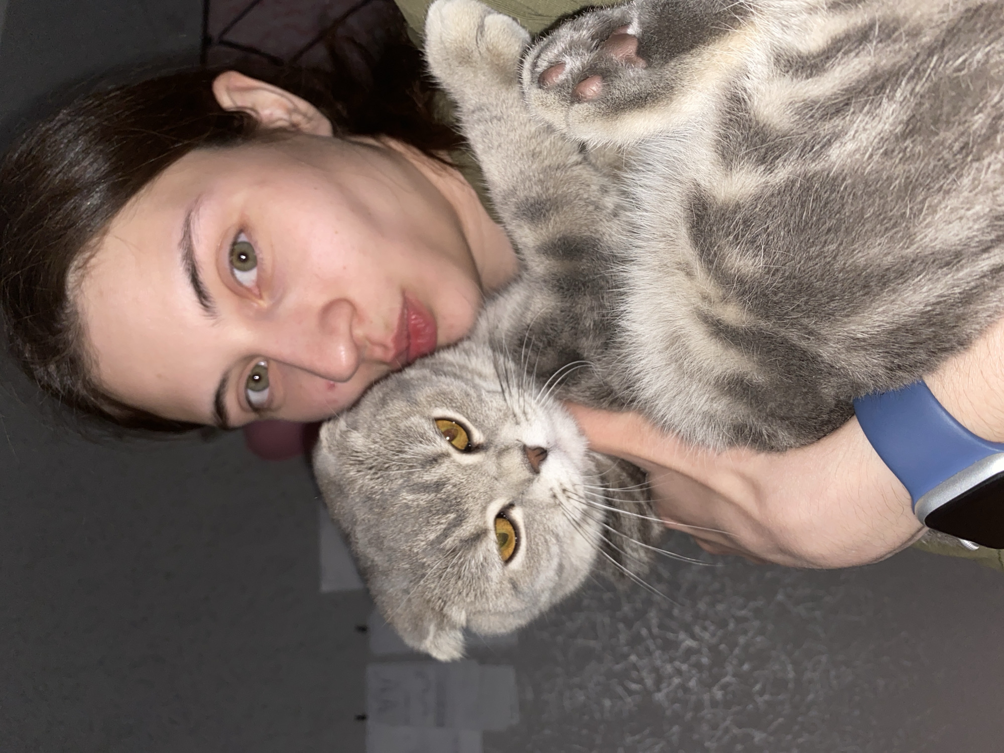 A young woman with dark hair and green eyes is pictured intimately with a gray tabby cat. She's holding the cat, its body across her arm, its head resting near her face. The cat has amber eyes and a flat-faced appearance, suggestive of a Scottish Fold or British Shorthair breed. The woman is wearing a blue smart watch on her wrist. The setting appears to be indoors, with a gray wall in the background. There's no evidence to suggest the image was taken in Votkinsk, Russia or any other specific location.