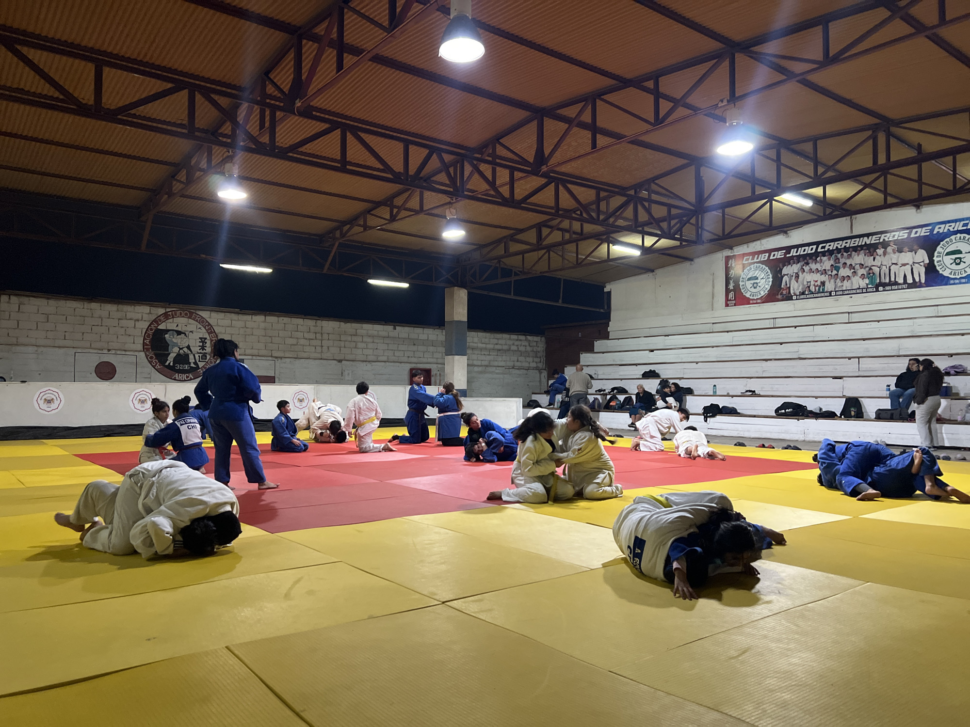 The image depicts a judo training session taking place indoors on a brightly lit mat. The mat is divided into red and yellow sections, common for judo practice. Several individuals, predominantly young people, are engaged in judo exercises and practice matches. Some are in the process of grappling, while others are in resting positions on the mat. In the background, spectators are seated on tiered bleachers, observing the training. A banner is visible on the wall behind the bleachers, advertising the "CLUB DE JUDO CARABINEROS DE ARICA," indicating the location as Arica, Chile. The setting appears to be a sports gymnasium or dojo, characterized by the high, industrial-style ceiling with exposed metal beams and corrugated roofing. Overhead lights illuminate the training area.