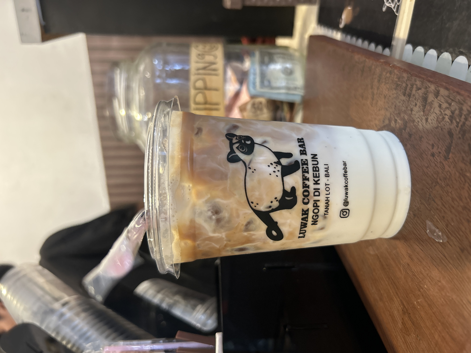 The image shows an iced coffee drink in a clear plastic cup, with a logo featuring a cat. The text on the cup reads "LUWAK COFFEE BAR NGOSI DI KEBUN TANAH LOT - BALI @luwakcoffeebar". The drink appears to be a latte or similar coffee beverage with milk and ice. The cup is placed on a wooden surface. In the background, there is a glass jar with money inside, and a blurred figure of a person in black clothing. The overall setting suggests a cafe or coffee shop. There is no information in the image to determine the location in the city of Kediri, Indonesia.