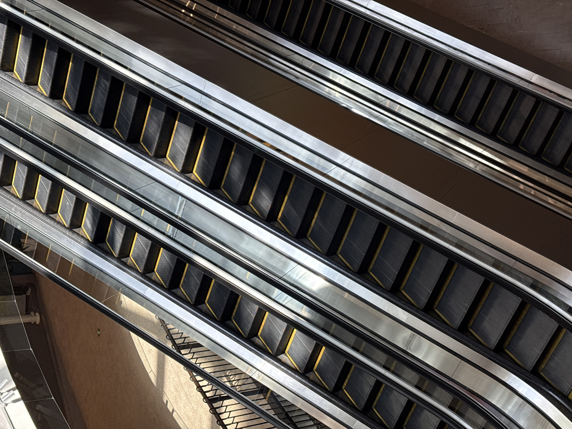 The image shows a top-down view of multiple escalators in a modern building, likely a shopping mall or transit station. The escalators are made of dark gray treads with yellow safety strips and have polished metal handrails and sides. Sunlight casts shadows across the steps of the escalators, highlighting their textured surfaces. In the background, a floor with a patterned carpet or tile is visible, along with a dark railing. The scene is devoid of people, emphasizing the architecture and design of the escalators themselves. The environment appears clean and well-lit, with a focus on the geometric lines and reflections created by the metallic surfaces. The precise location in Shanghai, China, cannot be determined from the image alone.