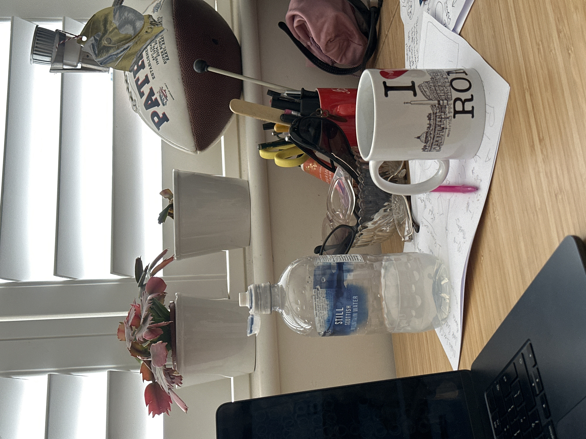 This is an indoor scene on a desk adjacent to a window covered with white blinds. The desk is cluttered with various items.
Objects on the desk include:
* A football with "PATR" visible, possibly a New England Patriots football.
* A desk lamp clipped onto the football.
* Three white potted plants with greenery.
* A red pencil cup with various stationery items.
* A white mug with "I ❤️ ROMA" printed on it, along with an image of Roman architecture.
* A bottle of "STILL SCOTTISH MOUNTAIN WATER" from M&S.
* A pair of sunglasses.
* Some papers with handwriting.
* A laptop, with the screen and keyboard visible.
The location in Tunbridge Wells, United Kingdom is unknown. The image does not depict any people, actions, or interactions.