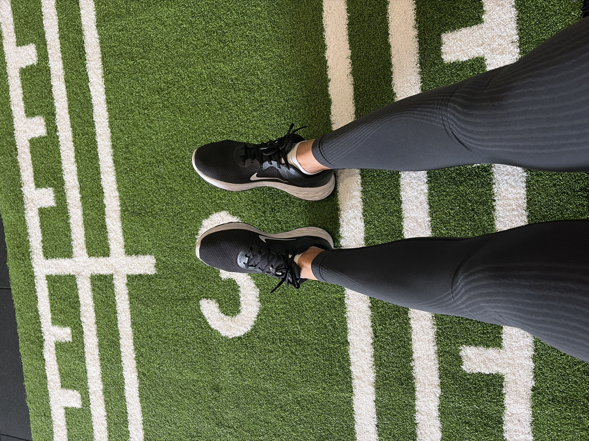 The image shows a person's legs and feet from a top-down perspective, standing on a green artificial turf surface with thick white stripes. The person is wearing black athletic leggings with a subtle textured pattern and black Nike running shoes with white soles. White socks are visible above the shoes. The white stripes on the turf appear to spell out "FIT" vertically. The lighting suggests an indoor setting, possibly a gym or fitness studio. There are no clear indicators of time of day or weather conditions. The overall impression is of someone ready for a workout or having just completed one. The scene is set in Tsarevo, Bulgaria.