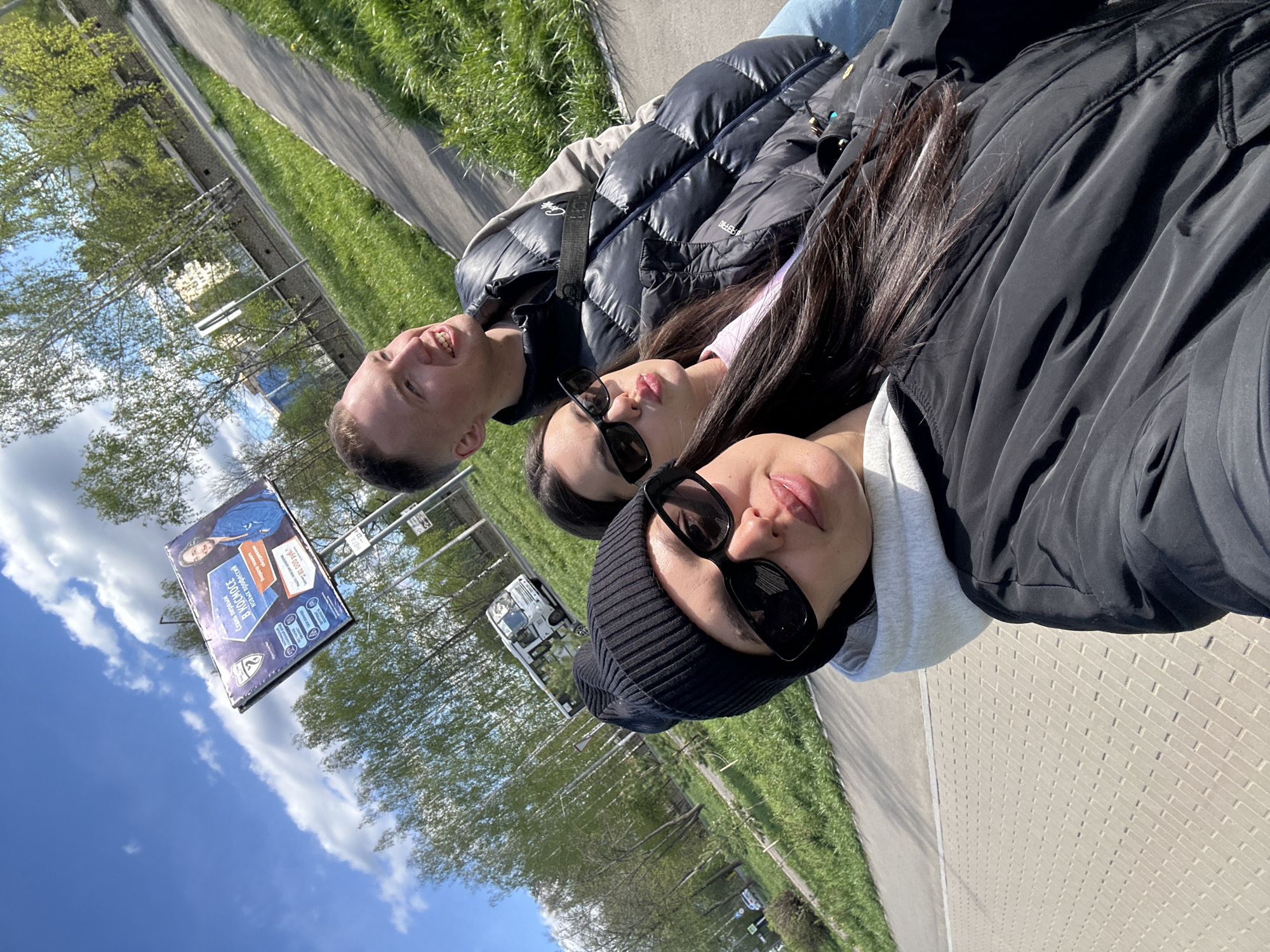 Three individuals are captured in a selfie taken outdoors in Chaikovsky, Russia. The person on the left is male, with short hair, wearing a black puffer jacket. The middle person is female, with dark hair and sunglasses, wearing a light-colored top under a black puffer jacket. The person on the right is female, wearing a black beanie, sunglasses, and a black jacket over a gray hoodie. They are standing on a brick-paved walkway, with a grassy area and trees to the right. To the left, there is a roadside and a billboard with Russian text. The sky is blue with scattered clouds. A white van is visible in the background.