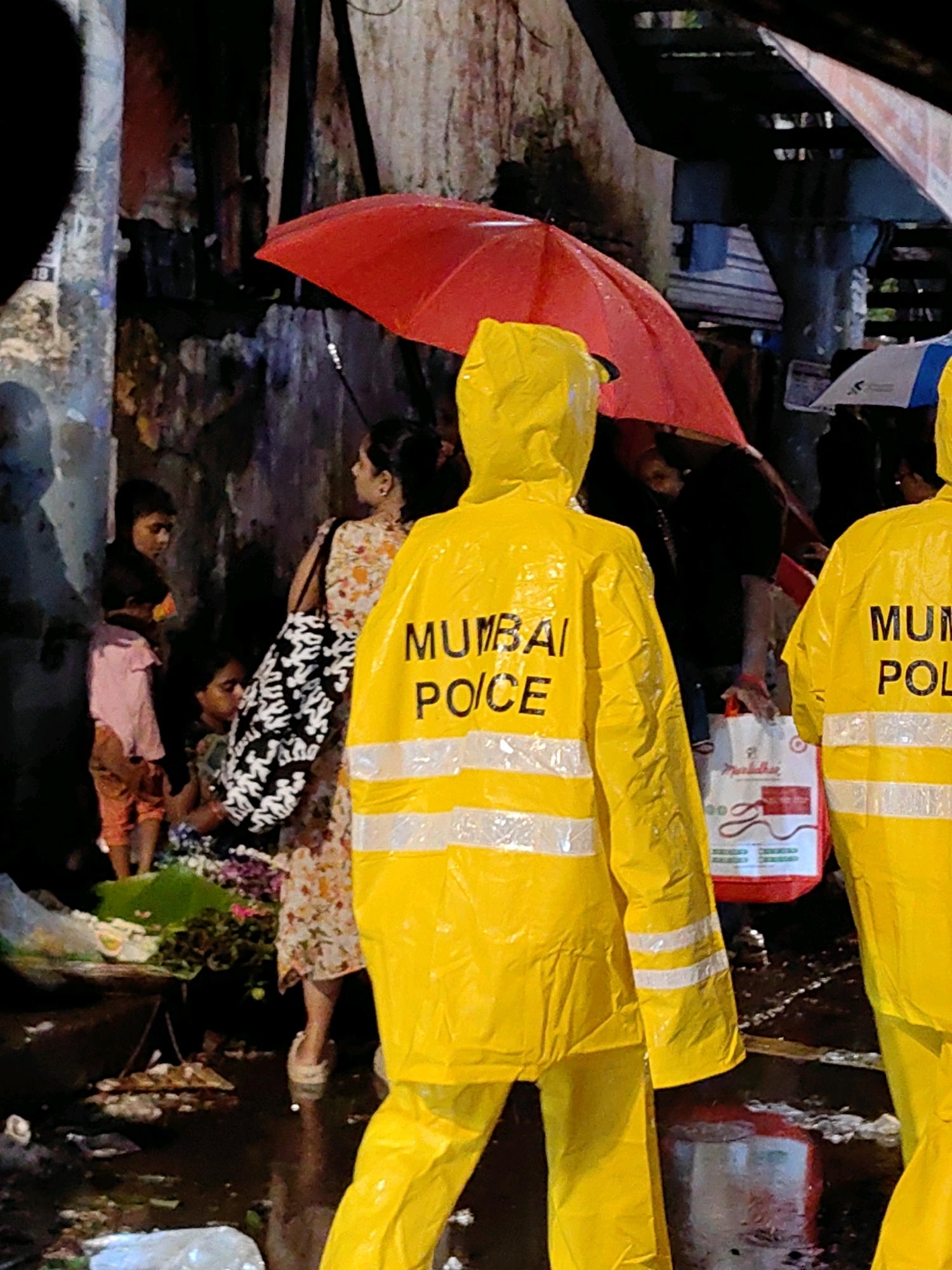 The image depicts a street scene in Mumbai, India, during heavy rain. Two individuals are prominently visible wearing bright yellow raincoats with reflective white stripes and the words "MUMBAI POLICE" printed on the back. The person on the left is facing away from the camera, while the person on the right is partially visible from the side. A red umbrella is held by someone in the background, and another umbrella, white and blue, is partially visible. Several other people, including children, are gathered in the scene. One woman in a floral dress carries a black and white patterned bag. The environment appears to be a market street, with wet ground, debris, and what looks like flowers or produce laid out on the wet surface. The lighting suggests it is either nighttime or a very overcast day. The presence of the "MUMBAI POLICE" raincoats indicates an official presence in response to the weather conditions or a related incident.