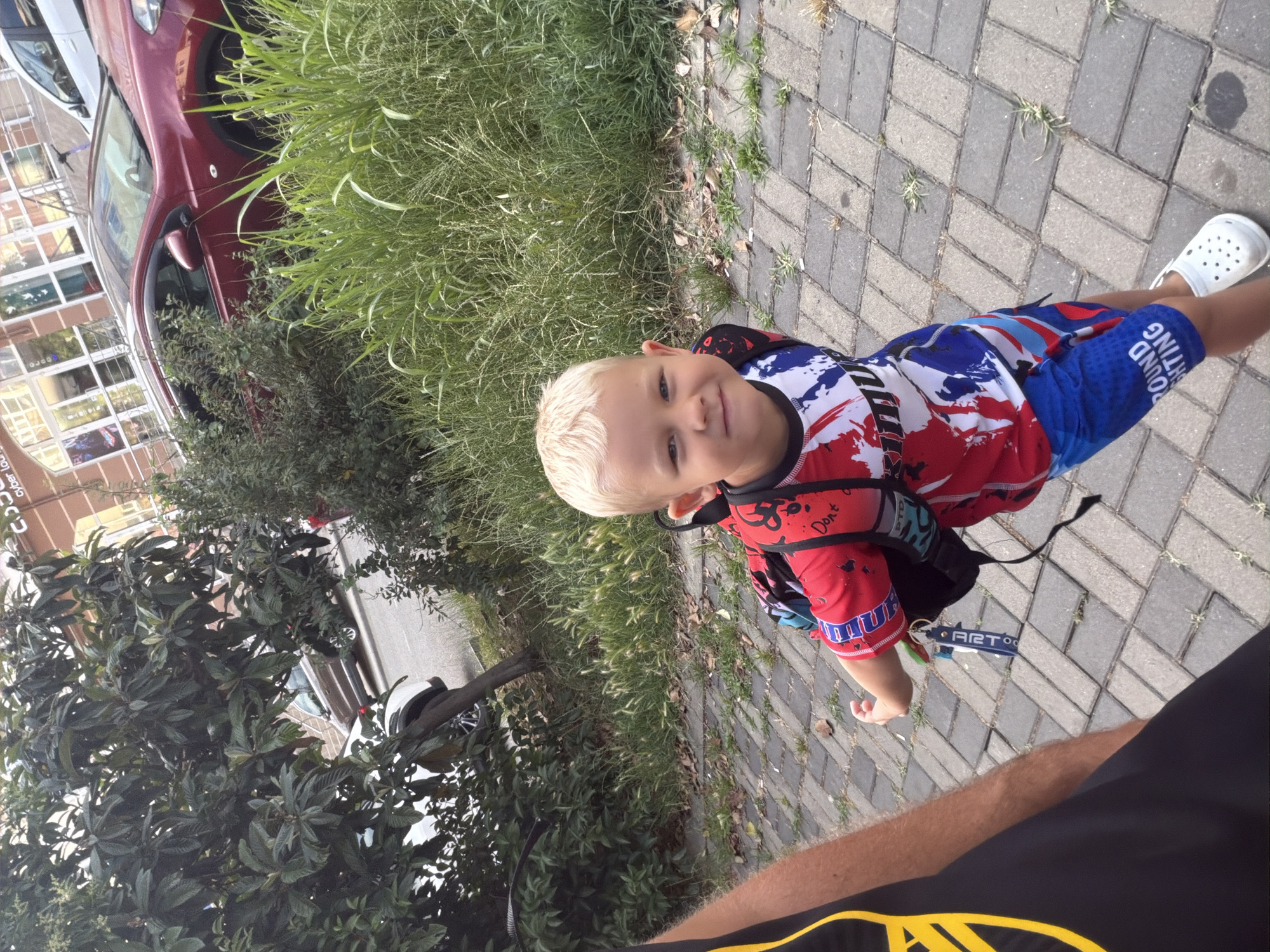 An outdoor, eye-level, full shot captures a young boy with blond hair and a bright smile. He is wearing a red, white, and blue shirt with a splatter paint design and blue patterned shorts. The boy is walking on a paved walkway made of grey and beige rectangular bricks. He has a backpack on his back with a green and blue keychain attached to the strap. To the left of the frame, the arm and shoulder of an adult are visible, wearing a black t-shirt with a yellow design. The background shows a street with cars, including a red hatchback and a white car, parked along the side. Green foliage and tall grass are present on the right side of the image, with trees on the left. The overall setting appears to be a residential or commercial area in the city of Sochi, Russia, possibly near a building with large windows and a sign that says "Cyber Lounge".