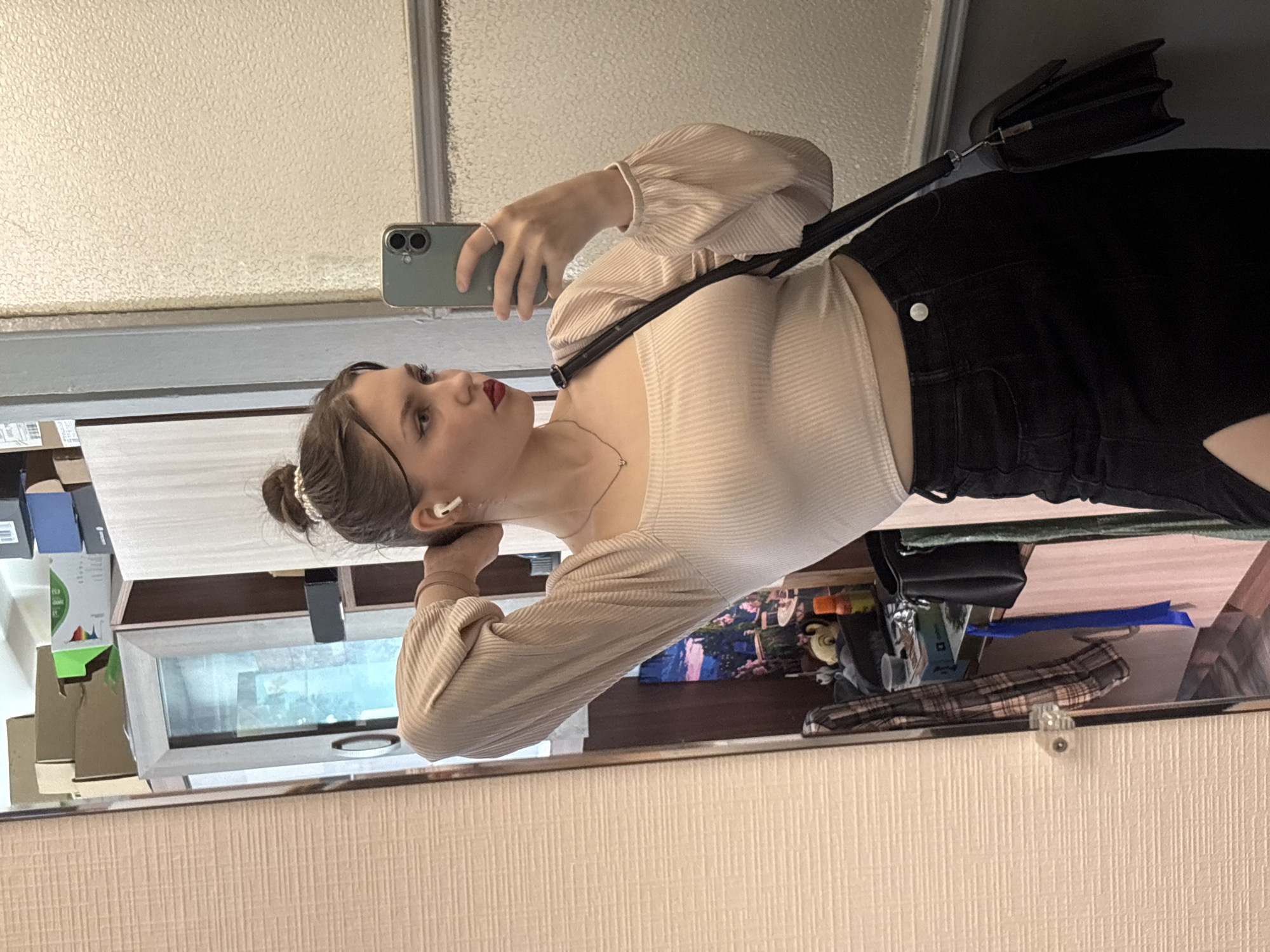 A young adult female is depicted taking a mirror selfie indoors in Saint Petersburg, Russia. She stands slightly angled, with her right arm raised behind her head, adjusting her dark brown hair which is styled in a high bun secured with a white beaded hair tie. Her left hand holds a dark green smartphone (appearing to be an iPhone with dual cameras), angled towards a large, rectangular mirror with a narrow, light-colored frame.
The female wears a light beige, ribbed crop top with a square neckline and puffed, long sleeves with elasticated cuffs. Her bottom attire consists of a high-waisted black denim skirt featuring a single silver button and a thigh-high slit on the left side. She has dark eyebrows, red lipstick, winged eyeliner, and wears white wireless earbuds in both ears. A thin gold-colored necklace with a small pendant is visible around her neck. A black shoulder bag with a long strap is slung across her body, resting on her right hip.
The mirror reflects a cluttered interior space. On the left, a light-colored wall with a horizontal textured pattern is visible. In the background, various cardboard boxes (including one with green accents and another with blue elements) are stacked on top of a light-colored cabinet or shelving unit. Below this, a window reflects an outdoor view of blue sky. A surface below the window displays a framed print of a landscape with pink flowers, an orange bottle with a white cap, a small yellow toy, a blue strap, and a plaid garment hanging. The direct wall surface where the subject stands, and a portion reflected to her right, is an off-white, textured material with some visible wear and a horizontal molding strip.