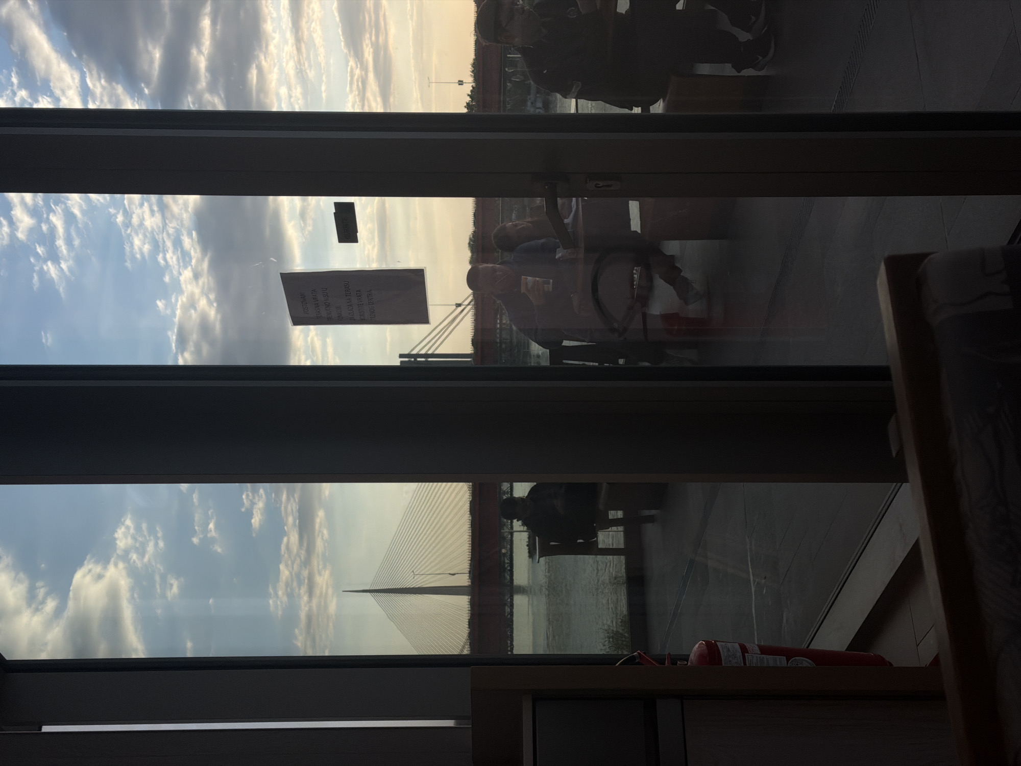 The image, originally captured in landscape orientation but presented here rotated 90 degrees clockwise, depicts a view from an indoor space looking out through a large window. The scene is a composite of reflections from the interior and an outdoor panorama, featuring the Ada Bridge over the Sava River in Belgrade, Serbia.
In the foreground, the windowpane reflects several individuals and interior elements. One person is distinctly visible sitting at a table, possibly holding a phone or small object. Other reflections suggest more people, both seated and standing, inside what appears to be a cafe or restaurant. A red fire extinguisher rests on a wooden ledge in the lower right portion of the indoor view.
Beyond the reflections, the prominent structure of a modern white cable-stayed bridge (the Ada Bridge) spans across the wide expanse of the Sava River. The sky is partially cloudy, illuminated by a warm, soft light, characteristic of late afternoon or early evening. This light highlights the clouds and the intricate cables of the bridge.
Text is visible on a sign reflected in the window. The Serbian phrases include "POSTOVANI" (Respected/Dear), "IZLAZ NA TERASU NIJE U FUMKCIJI" (Exit to the terrace is not in function/out of order), "ZATVORENA VRATA" (Closed door), and "KORISTITI ULAZ DESNO ENTRA" (Use the entrance on the right / Right entrance).
The image captures a moment of quiet observation from a comfortable indoor setting, offering a scenic view of Belgrade's cityscape at what appears to be the golden hour.