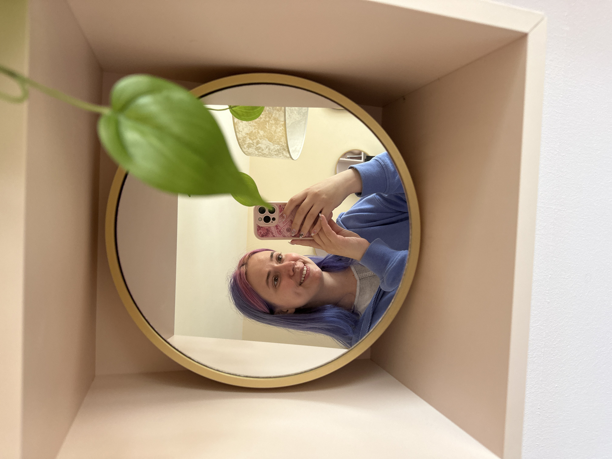 A young woman with blue and pink dyed hair is reflected in a round gold-framed mirror. She is smiling and holding a pink patterned smartphone in her hands, presumably taking a selfie. She is wearing a blue hoodie. The mirror is positioned in a shallow, peach-colored alcove or shelf unit against a white wall. A green plant leaf partially obscures the upper left corner of the mirror. In the mirror, a glimpse of a hanging lamp can be seen. The setting appears to be indoors. The location is not specified, so it can't be said that the image was taken in the city of Gijón/Xixón, Spain.