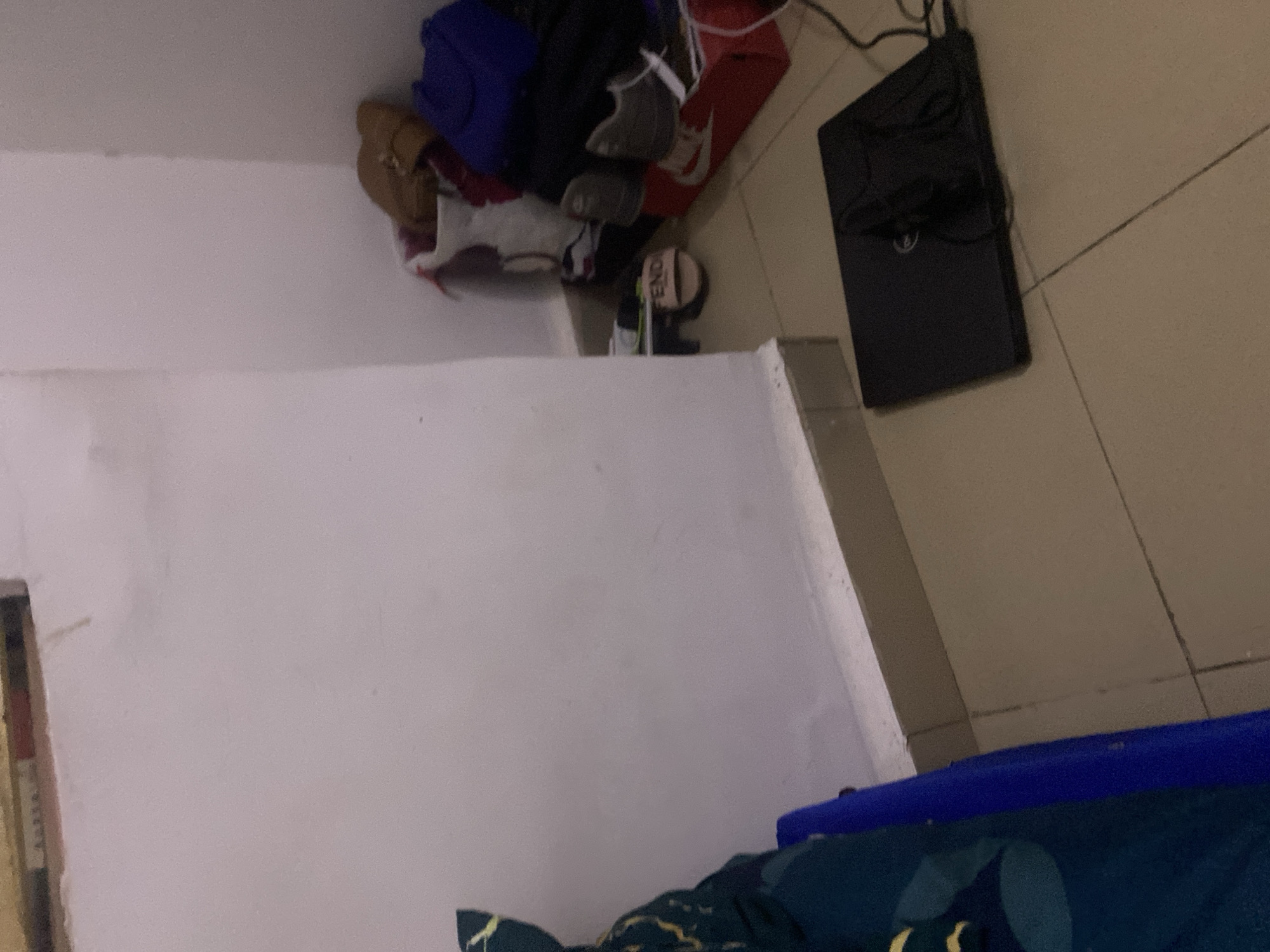 The image depicts a cluttered corner of a room. On the floor, a black laptop is positioned. To the right, a collection of items is piled near the wall, including shoes, bags, and what appears to be clothing. One pair of footwear, a slide with the word "FENDI" visible, rests on the floor. The walls are painted a light color, and the floor is tiled in a neutral tone. There are no people visible in the image. The scene suggests a lived-in space with personal belongings. No specific details about the city of Howayi, Nigeria, can be inferred from the provided visual information.