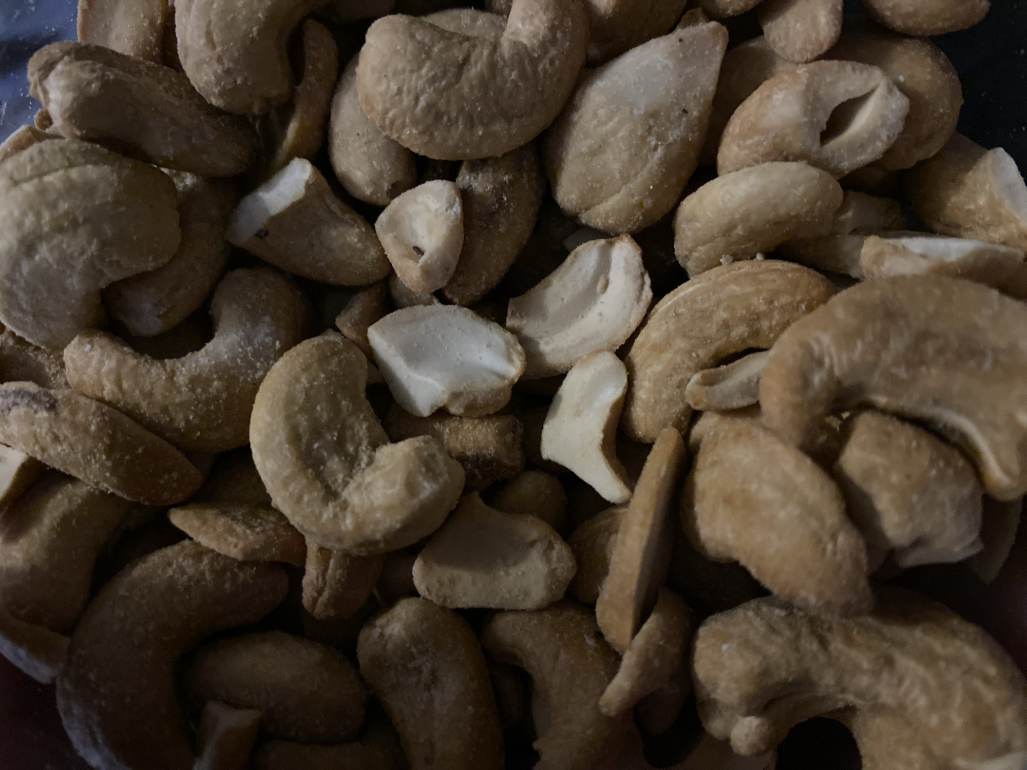 The image shows a close-up of a pile of cashews. The cashews are a light brown color and appear to be salted. Some of the cashews are whole, while others are broken into pieces. The background is dark and out of focus. There is no indication of location in Konstantinovo, Bulgaria or any information about people, actions, interactions, or the environment.