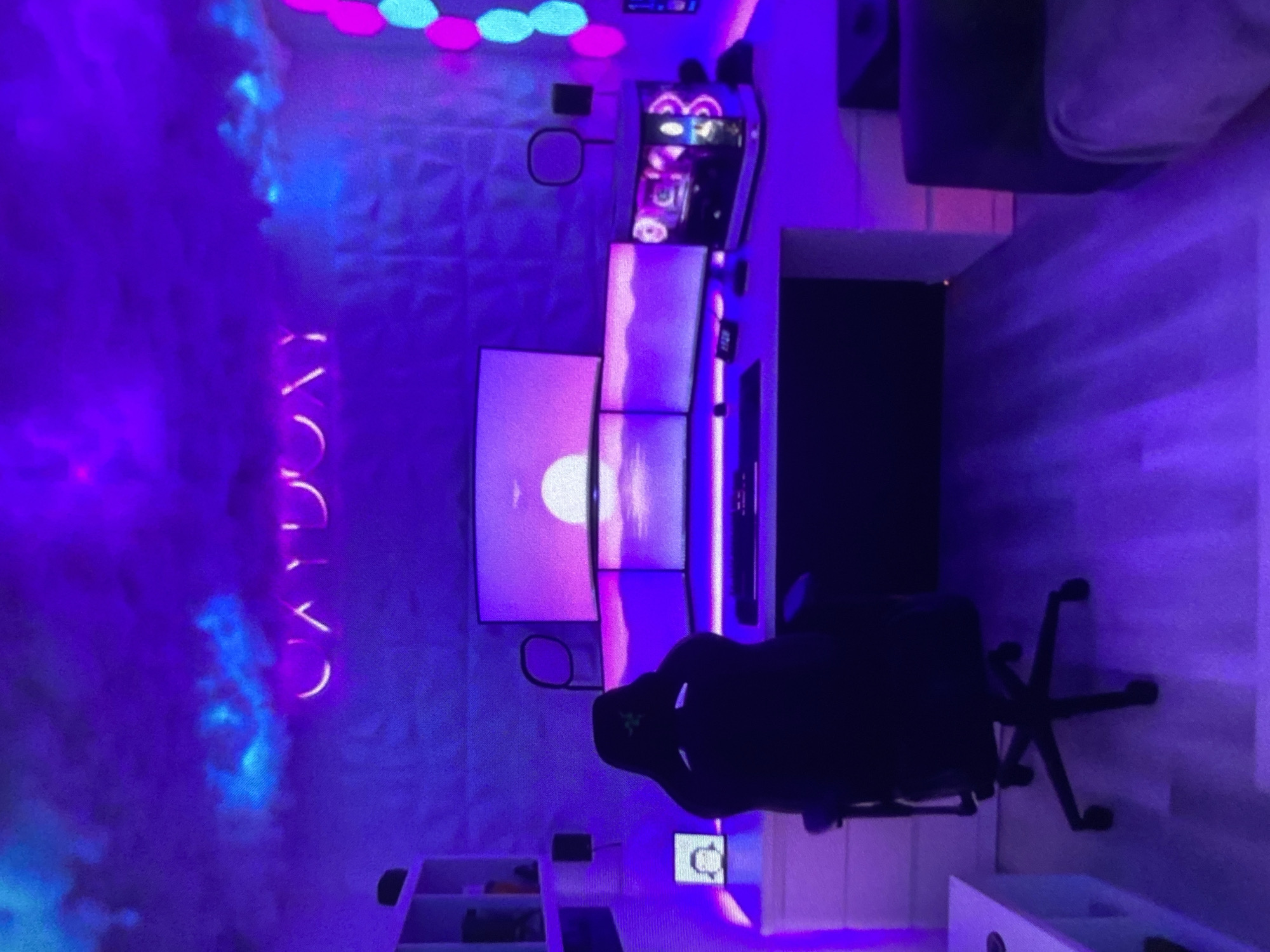 The image depicts a gaming setup in a room with purple and blue lighting. A multi-monitor display is visible on a white desk, with a gaming chair in front of it. The room features geometric wall panels and illuminated shapes. To the side, there is a shelving unit containing items. The image does not provide sufficient information to determine the location within the city of Lhokseumawe, Indonesia. There are no people visible in the image.