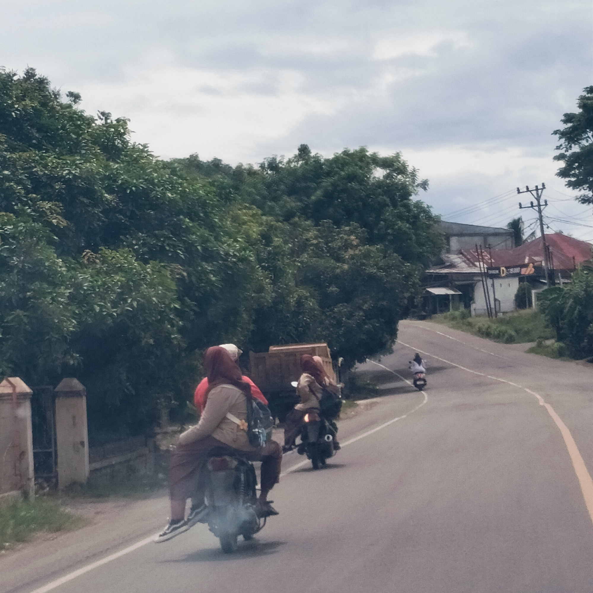 The image depicts a scene on a paved road in Blangdalam, Indonesia, under an overcast sky. The view is from behind, looking down a road that curves gently to the right, bordered by dense green trees and vegetation on both sides.
Several individuals are seen riding motorcycles or scooters. In the immediate foreground, two people are on a single scooter, possibly an older model, which is emitting exhaust smoke. The driver wears a light brown top, dark pants, and a dark red/maroon hijab, carrying a backpack. The passenger, also in a similar hijab, is partially visible. Behind them, another individual on a more modern scooter, similarly dressed, follows. Further down the road, a third person is riding a smaller moped.
On the right side of the road in the distance, several houses with red roofs and light-colored walls are visible through the trees. A wooden utility pole carrying power lines stands near the houses. On the left side, close to the viewer, two concrete fence or gate posts are partly visible. The sky is cloudy with light grey clouds. A white sign with dark, unreadable text is faintly visible on one of the buildings in the background. The road features a solid white line on the left and a yellow line on the right edge. The scene captures everyday travel in a green, semi-rural setting.