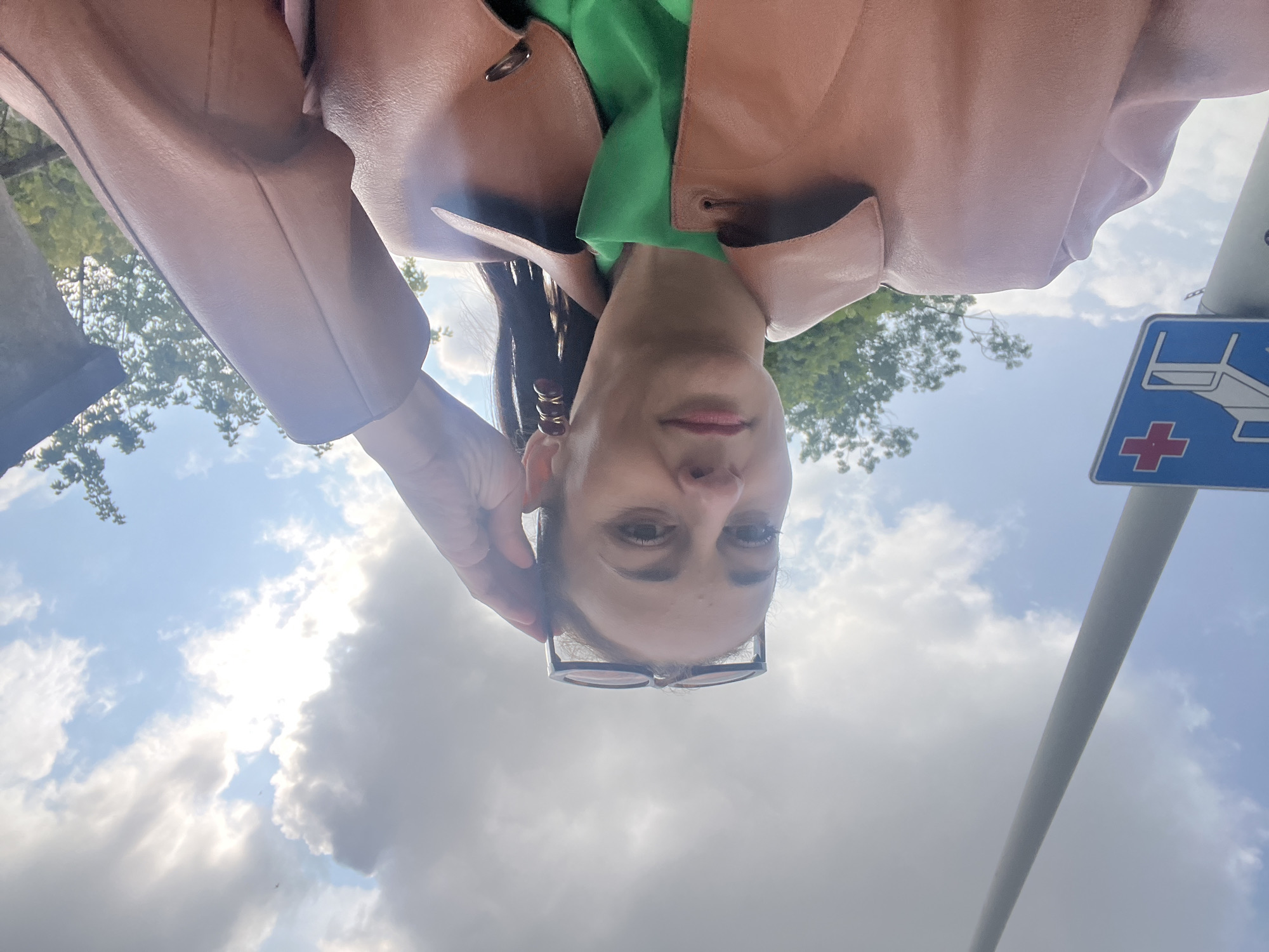 A woman is taking a selfie outdoors. She is wearing a brown leather jacket over a green top and has sunglasses perched on her head. Her right hand is raised, touching her hair. Behind her, a blue sign with a red cross and an image of a medical cot is visible, attached to a pole. The sky is cloudy with patches of blue. Trees are visible in the background.