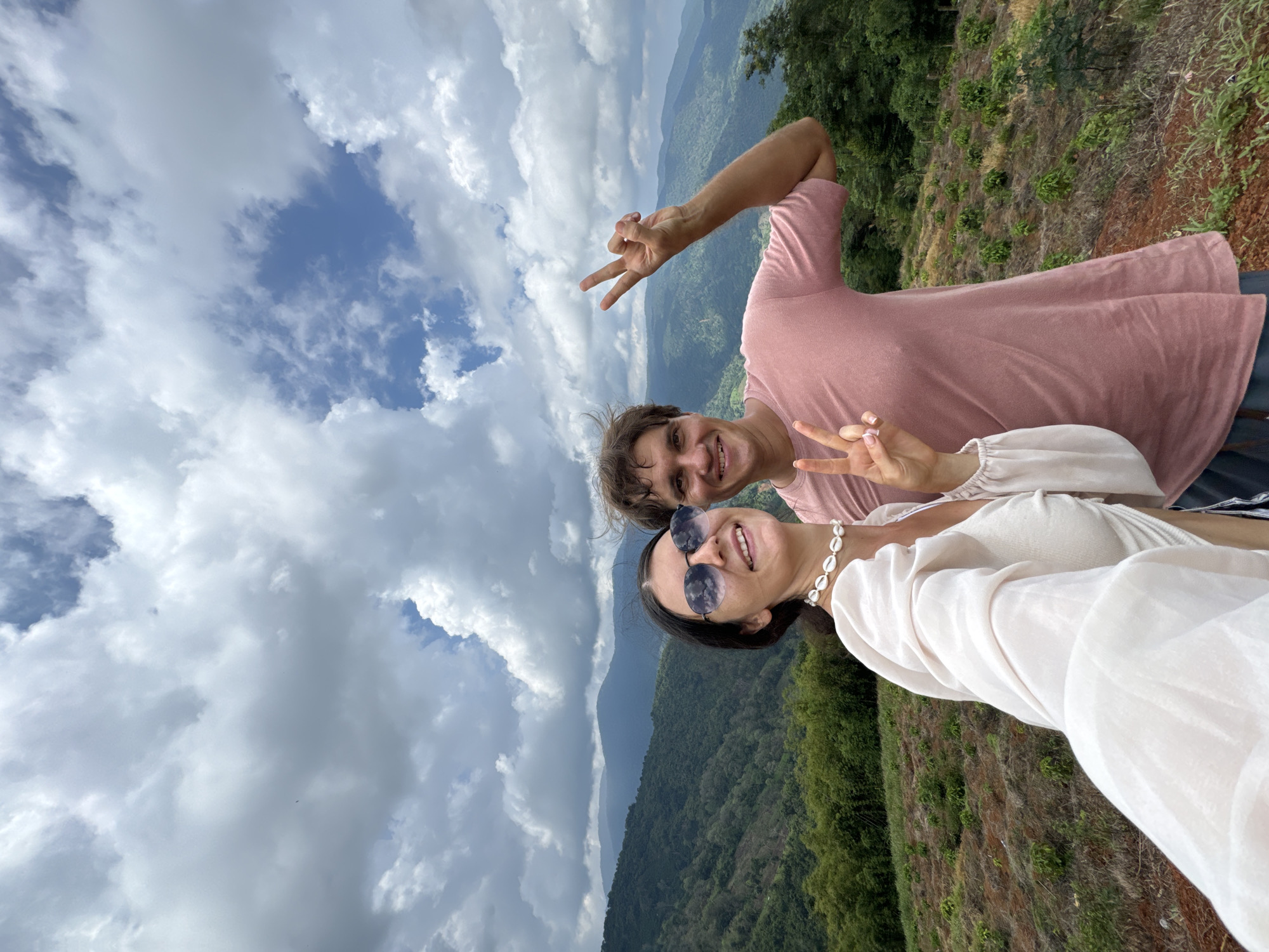 A man and a woman are posing for a selfie in a mountainous landscape with a cloudy sky. The woman, on the left, wears a white top and sunglasses, and holds up two fingers in a peace sign. The man, on the right, wears a pink t-shirt and also holds up two fingers in a peace sign. The background shows rolling green hills and mountains, indicative of a rural or natural setting. The location is identified as Nao Sẻ, Vietnam. The terrain in the foreground appears to be a cultivated field with some sparse vegetation. The overall mood is cheerful and relaxed.