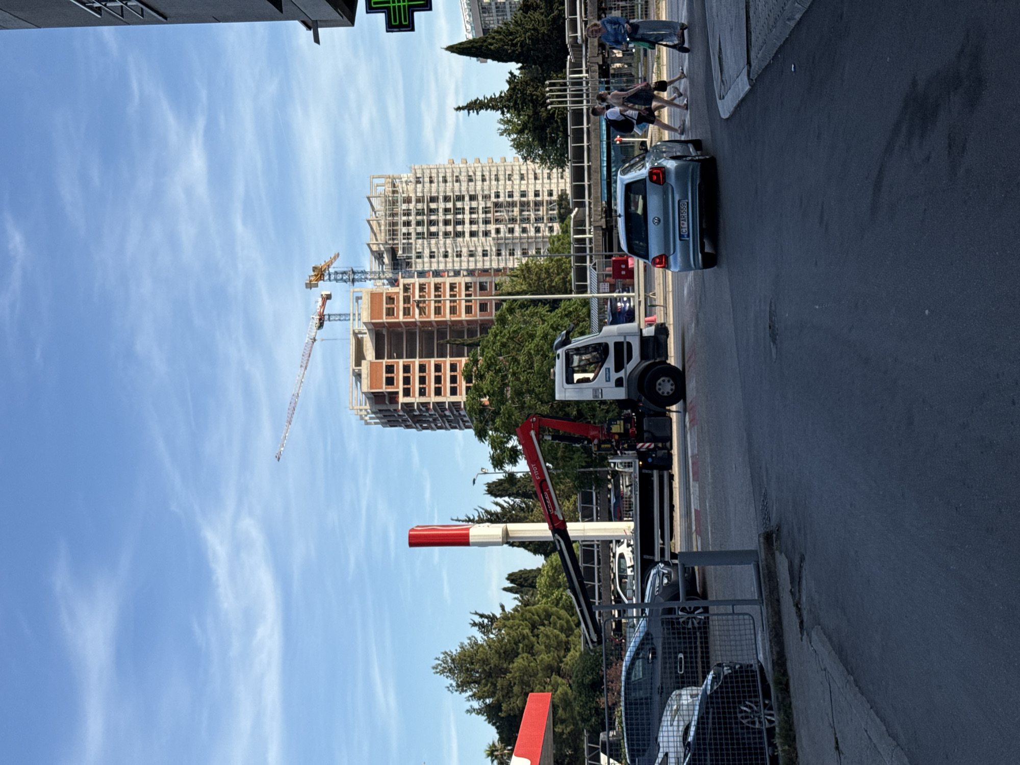 This urban scene in Budva, Montenegro, depicts a street with ongoing construction activities under a bright, clear blue sky with scattered white clouds.
In the background, multiple tall buildings are under construction. One building shows a skeletal concrete frame with brick infill, while another appears further along with a multi-story, grid-like facade. Several large construction cranes are visible, indicating active development.
On the street, pedestrians are present, with two women crossing the road in the mid-ground and another group walking on the sidewalk. A light blue Volkswagen Polo car with the license plate "CT AR559" is visible, either parked or moving slowly. Further down the street, a white truck equipped with a red FASSI crane is parked. Other cars are partially visible, some behind a metal fence. A red and white barrier arm, possibly controlling access to a parking area or construction site, stands prominently in the foreground. A green cross symbol, commonly associated with pharmacies, is visible on a sign or traffic light in the upper left corner. The image captures a lively daytime setting, typical of a growing city.