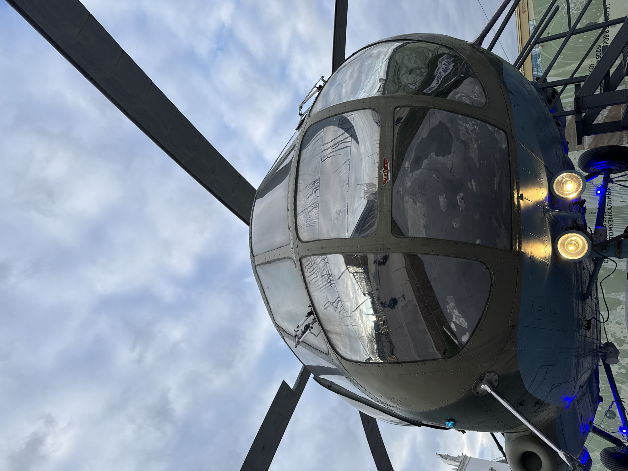 The image features a close-up, low-angle shot of a helicopter's cockpit, showcasing a complex arrangement of glass panels framed by a riveted, dark green and blue fuselage. The glass reflects the surrounding environment, including clouds and hints of buildings. The reflection reveals the architecture of the location. The helicopter's propeller blades extend from the central hub, reaching towards the cloudy sky. The landing gear is visible on the right side of the frame, illuminated by bright lights. The scene is in the city of Moscow, Russia.