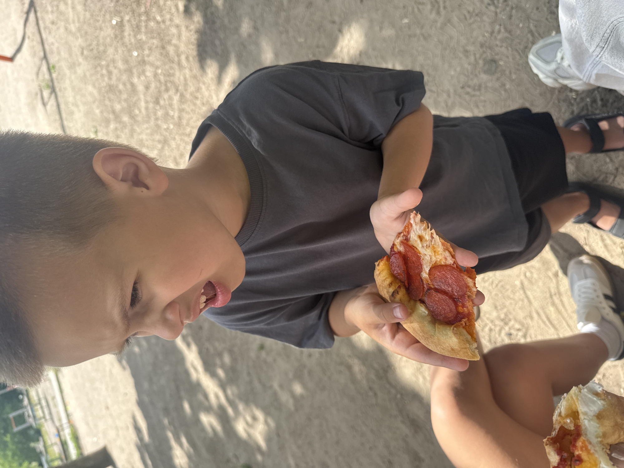A young boy with short brown hair and a grey t-shirt is holding a slice of pepperoni pizza and looking to his left with his mouth open. He is standing on a dirt path with scattered tree shadows. In the foreground, another slice of pizza is being held by an unseen person, along with a person's legs wearing white sneakers. In the background, there are glimpses of a playground and trees. The setting appears to be an outdoor park or recreational area. There is no specific indication of the location being the city of Dnipro, Ukraine.