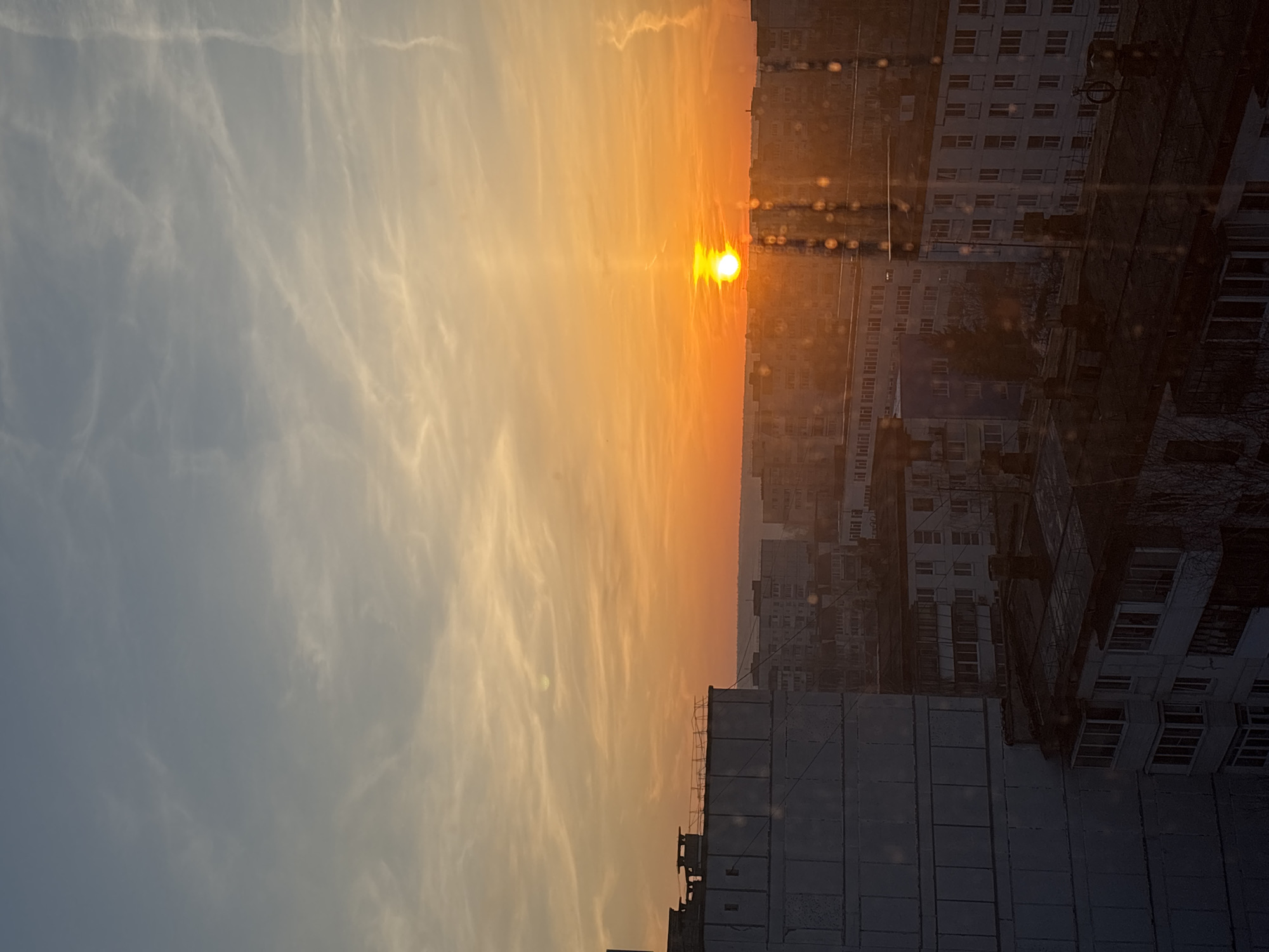 The image depicts a sunset in an urban setting, likely in Chaikovsky, Russia, given the prompt. The sun is positioned near the horizon, casting a bright orange glow. The sky transitions from a light blue with wispy clouds to an orange hue near the sunset. To the right of the image, there are multiple high-rise buildings, their windows visible, suggesting residential or commercial use. The lower part of the image shows the top of a large, grey building under construction or renovation. The overall impression is one of urban life at dusk. There are no people visible in the image.