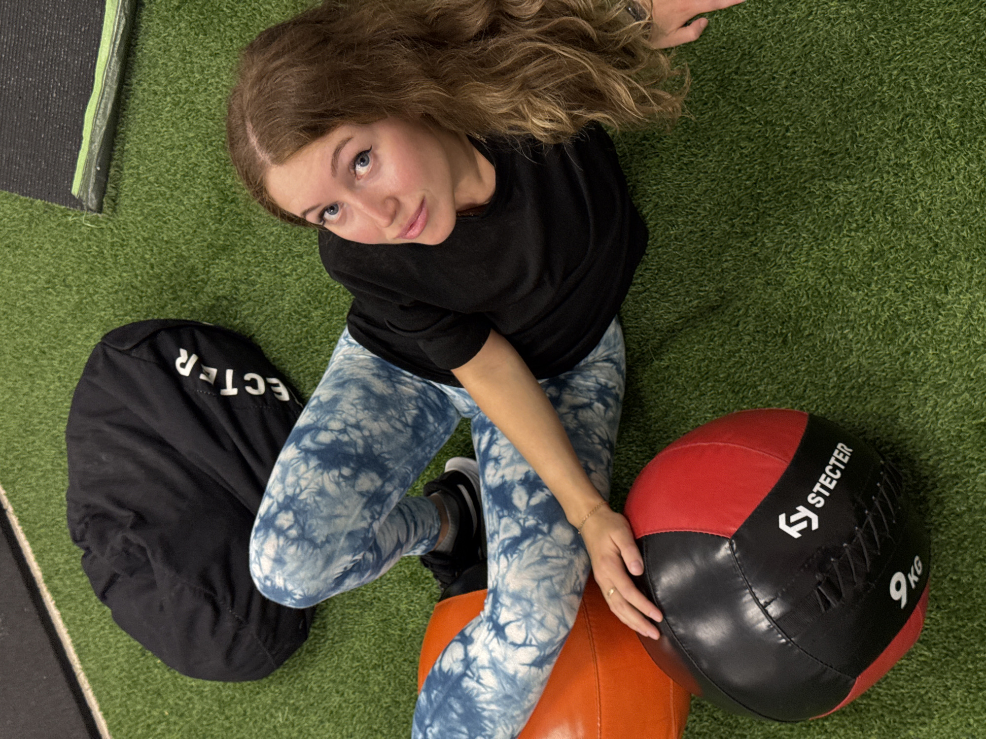 A young woman is depicted sitting on a green artificial turf floor, likely within a gym or fitness facility in Chaikovsky, Russia. She has long, wavy brown hair and blue eyes, looking upwards directly at the camera with a calm expression. She is dressed in a black t-shirt and blue and white tie-dye leggings. Her legs are positioned over an orange, possibly cylindrical or spherical, piece of gym equipment.
To her left, a black bag or sandbag is visible on the turf, with part of a white label reading "ECTER". To her right, a red and black medicine ball rests on the floor, displaying a white "STECTER" logo and the weight "9 KG". A black mat with a green stripe is partially visible in the upper-left corner of the frame. The setting appears to be indoors with bright, even lighting. The woman seems to be resting or posing for the camera during or after a workout.