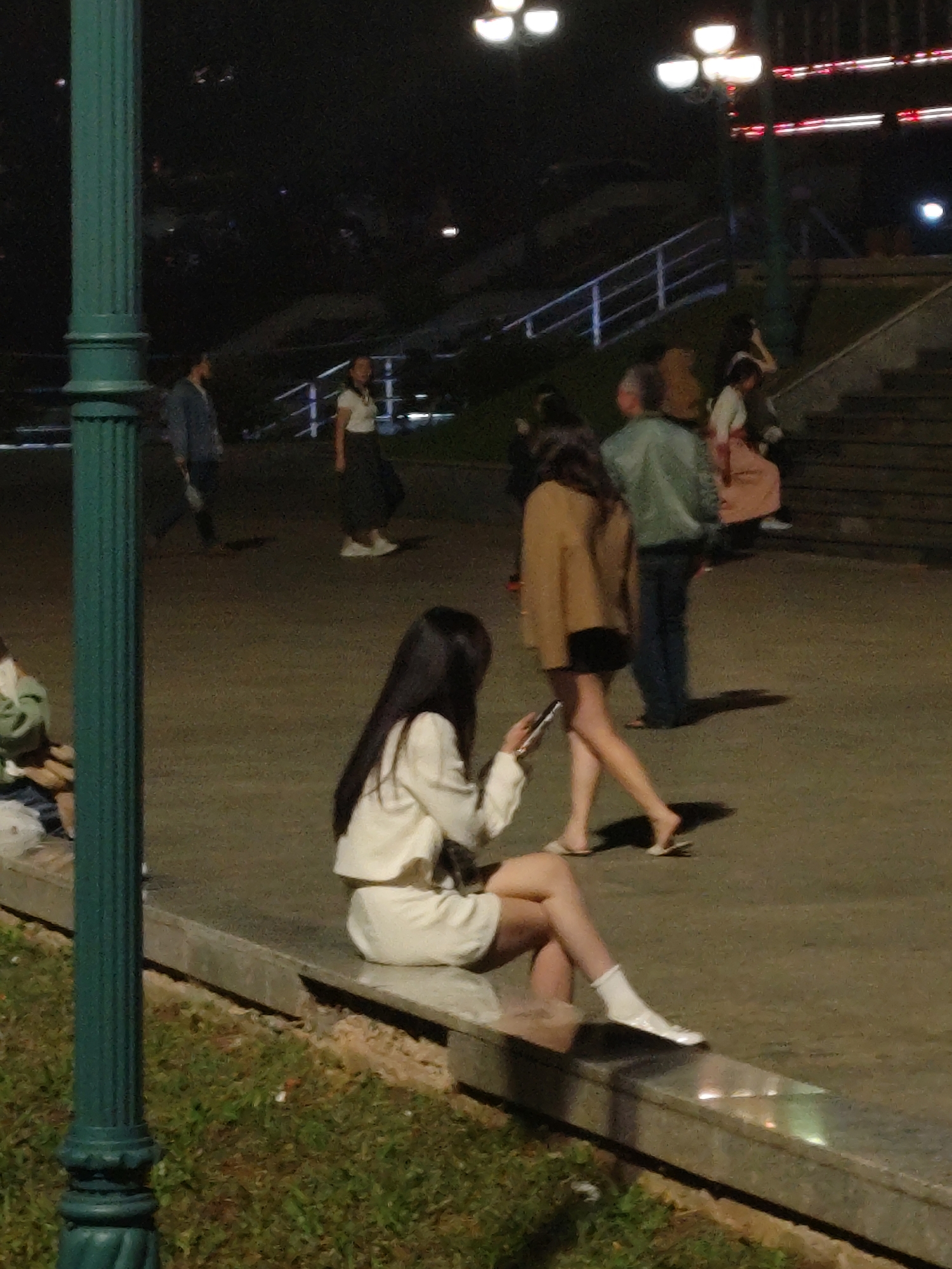 This media file depicts an outdoor scene at night, likely in Da Lat, Vietnam. The primary subjects are several people walking and sitting. In the foreground, a woman with long dark hair is seated on a concrete ledge, wearing a white outfit and white socks, looking down at her phone. Behind her, various individuals are visible. To the left, a person in a blue jacket walks away from the camera. In the mid-ground, a woman in a tan jacket and black shorts walks away, while a person in a light blue jacket and dark pants walks towards the right. Further back, more people are seen, including someone in a pink skirt and a white top.
The setting appears to be a public area with paved ground and grass. There are streetlights casting a warm glow, indicating it is nighttime. A dark green lamppost stands prominently in the left foreground. In the background, a set of stairs with railings and more lights can be seen, suggesting a park, plaza, or promenade. The weather appears to be clear, as there are no visible signs of rain or inclement conditions. The overall mood is casual and relaxed, with people engaging in everyday activities like walking and using their phones. No visible text is present in the image.