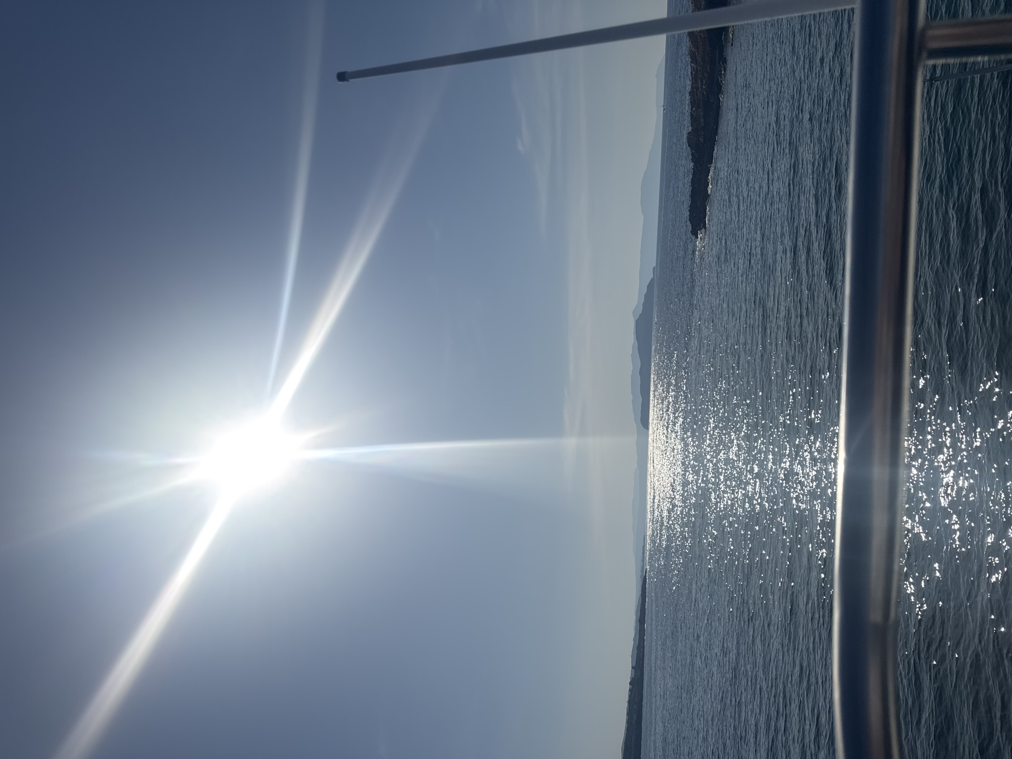 A bright sun shines intensely in a clear blue sky, casting a lens flare across the upper left quadrant of the image. Below, a wide expanse of dark blue sea sparkles with sunlight. In the distance, a hazy coastline with rolling hills is visible under the bright sky. In the foreground, a metallic railing runs horizontally across the bottom of the frame. A tall, white pole extends vertically along the right edge of the image. The scene is likely a view from a boat or a waterfront location in Chania, Greece, given the characteristic clear skies and coastal landscape of the region.