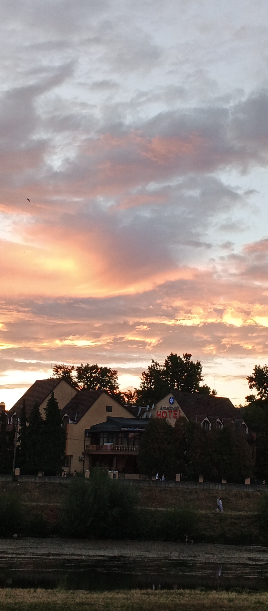 The image shows a sunset over Mukachevo, Ukraine. The sky is filled with colorful clouds, ranging from pink and orange to grey. In the middle ground, there is a hotel with a red "HOTEL" sign and a blue snowflake-like logo. Several buildings with brown roofs are visible, surrounded by trees. In the foreground, there is a body of water, possibly a river, with some greenery on the banks. A few people are scattered on the grassy embankment behind the water, and a bird is flying in the sky. The overall scene depicts a peaceful evening in a city setting.
