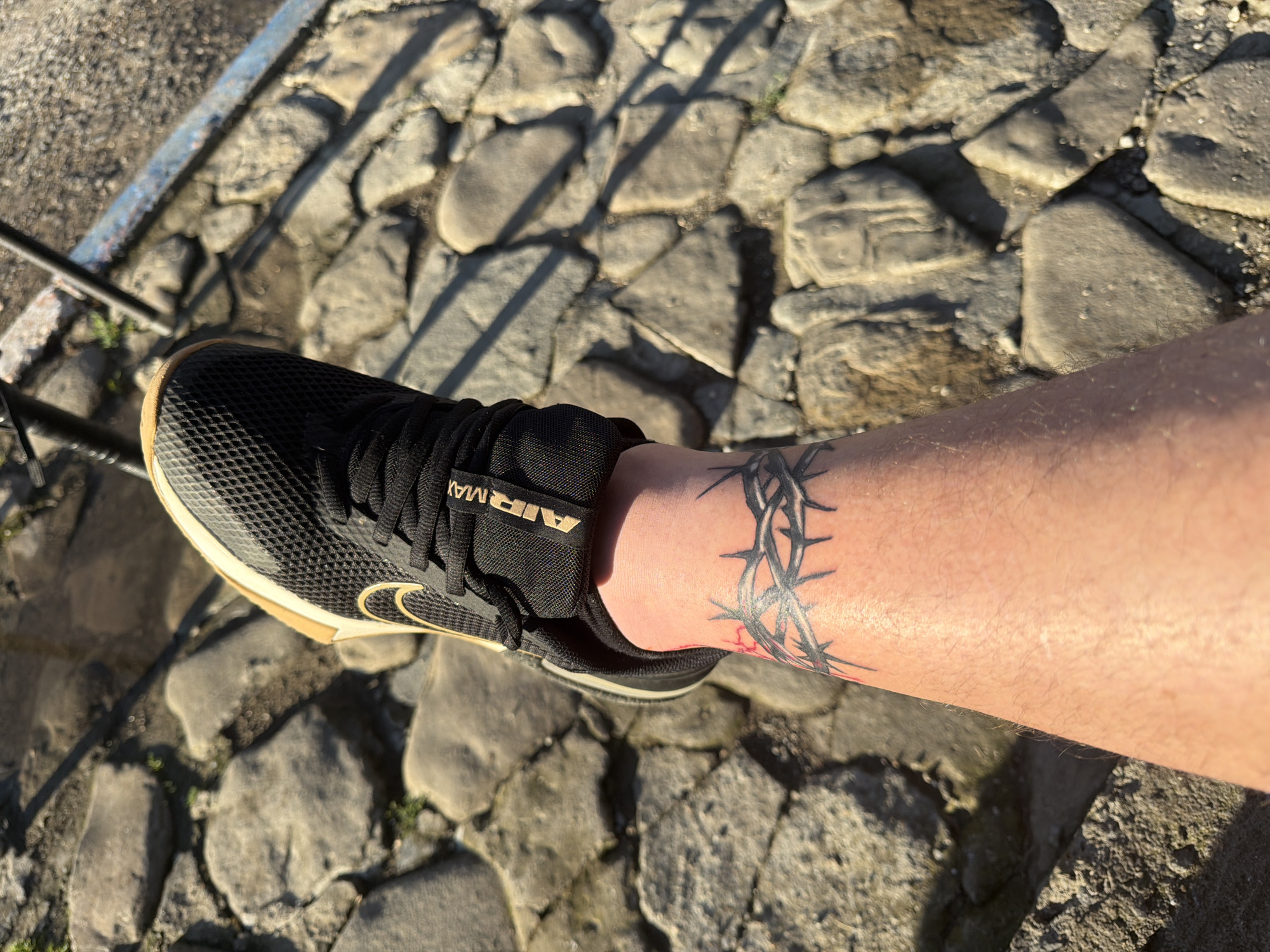 An overhead, close-up shot captures a person's lower right leg and foot resting on an outdoor paved surface in Shumen, Bulgaria. The foot is clad in a black Nike Air Max athletic shoe, which features gold-colored accents on the sole's edge and the iconic Nike swoosh logo on the side. The "AIR MAX" branding is visible on the shoe's tongue, oriented upside down relative to the viewer.
A prominent tattoo encircles the ankle, depicting a band of thorny branches, predominantly rendered in shades of dark grey and black with subtle touches of red, possibly suggesting blood or specific detailing. The ground beneath is composed of irregularly shaped, natural-looking grey and brown stones, giving the surface a rugged, cobblestone-like appearance.
The scene is brightly illuminated by direct sunlight, casting crisp, well-defined shadows from the shoe and leg onto the stony ground, indicating a clear, sunny day. A dark metallic railing or bar is partially visible in the upper left of the frame, hinting at the presence of a pathway, steps, or a protective barrier. No activity beyond standing is implied, and no emotions are discernible from this specific view.
