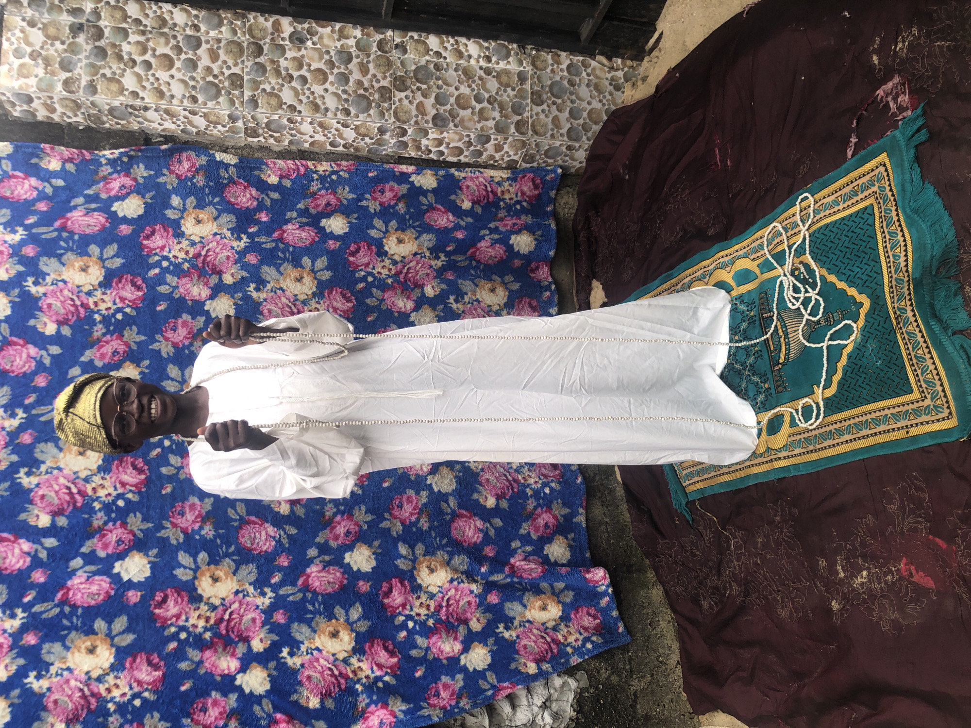 A full-body shot depicts a Black boy smiling broadly while standing on a prayer rug in front of a floral-patterned blue cloth. He is wearing a white thobe and a gold kufi hat, and he holds a string of white prayer beads. The prayer rug, which is teal and gold, is laid out on a dark maroon cloth, which is on the ground in an outdoor setting. The background consists of the floral cloth, a wall with circular patterned tiles, and a dark metal gate. The setting appears to be a residential area in Ado, Nigeria.