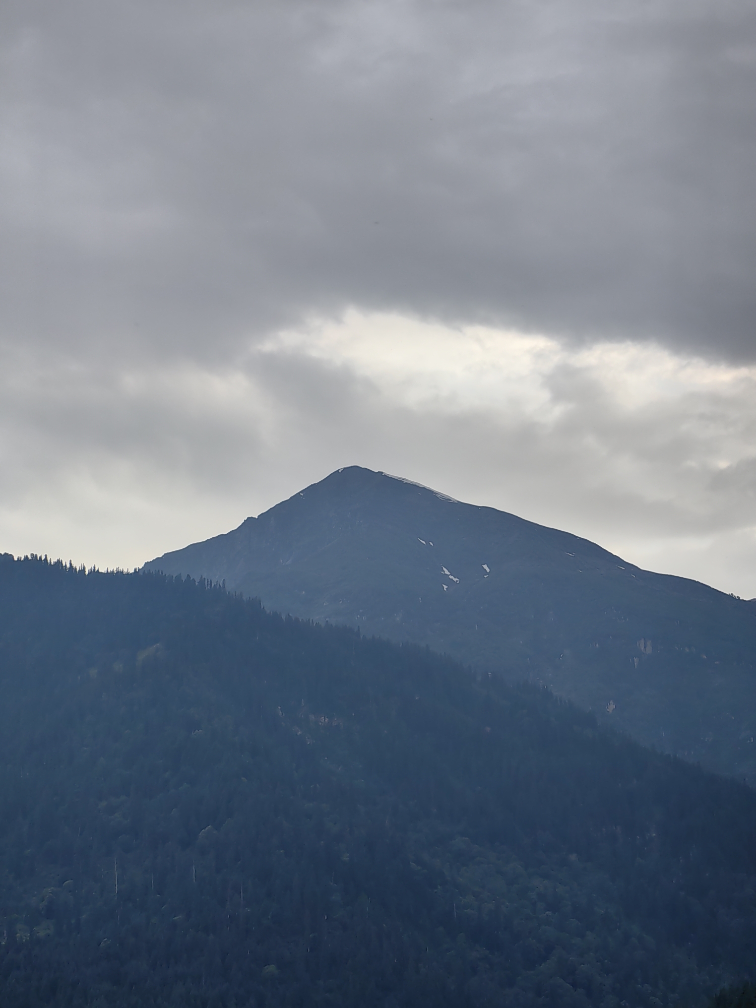 This media file features a serene landscape of a majestic mountain peak rising above a dense, dark green forest. The sky above is overcast with heavy, gray clouds, suggesting a gloomy or potentially rainy day. Hints of snow can be seen on the upper slopes of the mountain, indicating a high altitude. The foreground is dominated by a vast expanse of trees, their dark hues creating a stark contrast with the lighter tones of the mountain and sky. The overall mood is one of quiet solitude and natural grandeur. The scene appears to be captured during daylight hours, although the cloudy conditions obscure the direct sunlight. There is no text visible in the image, and no people or specific objects are depicted, focusing solely on the natural elements of the mountain and forest. Given the location context of Vashisht, India, this likely depicts a view of the surrounding Himalayan foothills.