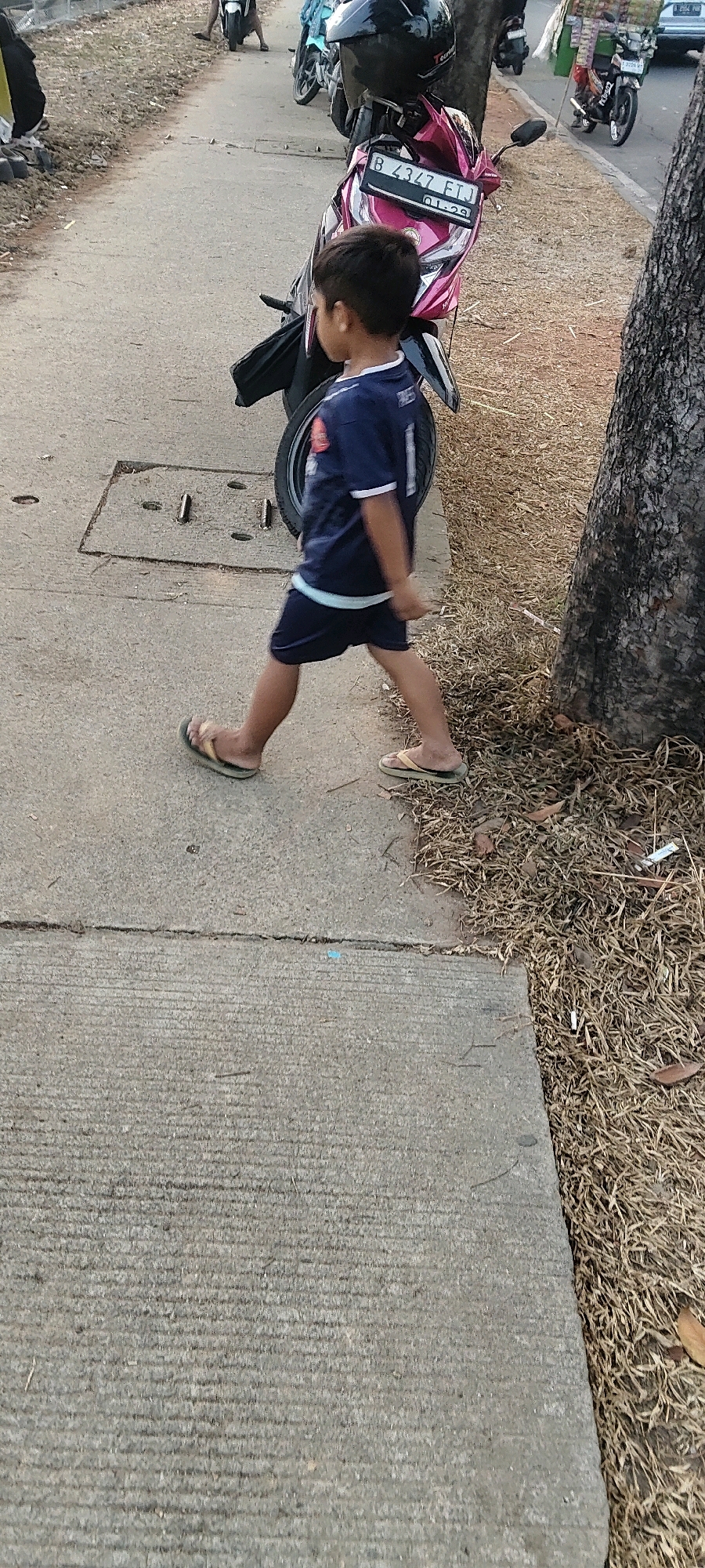 A young boy wearing a dark blue t-shirt with white trim and dark shorts is walking on a concrete sidewalk next to a tree. He is wearing flip-flops. Behind him is a pink and black motorcycle with a license plate that reads "B 4347 FTJ". The environment appears to be an outdoor setting, possibly in Pasirsari, Indonesia, with dry grass and dirt on the right side of the frame. Other motorcycles and people are visible in the background on a paved road.