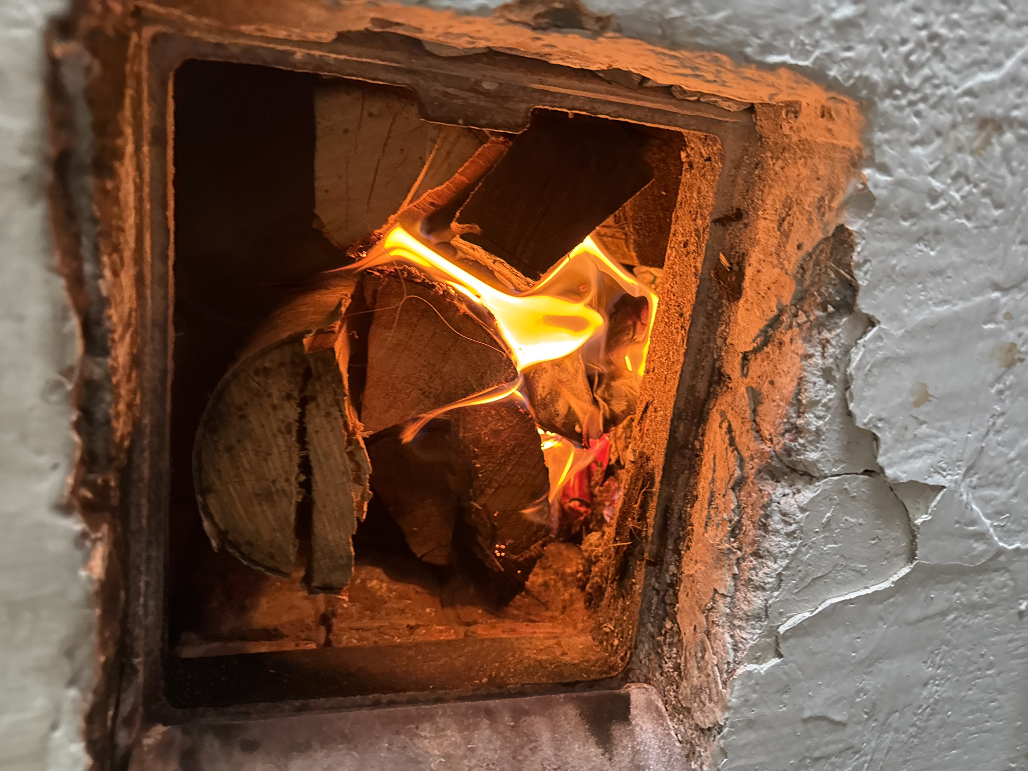 The image shows a fire burning inside a small, square, brick-lined opening in a rough, textured wall. The wood logs are stacked inside, with flames engulfing them and casting a warm orange glow. The wall surrounding the opening is a mix of grey and textured brown, appearing weathered and possibly peeling. The scene is likely indoors, depicting a small fireplace or oven. There is no information in the image that it is in the city of Бородулино, Russia.