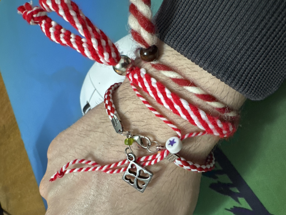 The image displays a close-up view of a human left forearm and wrist, situated indoors in Selanovtsi, Bulgaria. The wrist is adorned with multiple bracelets constructed from red and white twisted cords. One bracelet features a silver-colored four-leaf clover charm, a small green spherical bead, and a white cylindrical bead with a purple star, attached to a lobster clasp. Other bracelets include a thick braided red and white cord with a large silver-colored spherical bead, and another red and white twisted cord with a dark brown spherical bead. The lower right portion of the image shows the dark grey, ribbed cuff of a long-sleeved garment on the subject's arm. The background includes a bright blue surface, a white object resembling a computer mouse, and a green surface with indistinct patterns.
