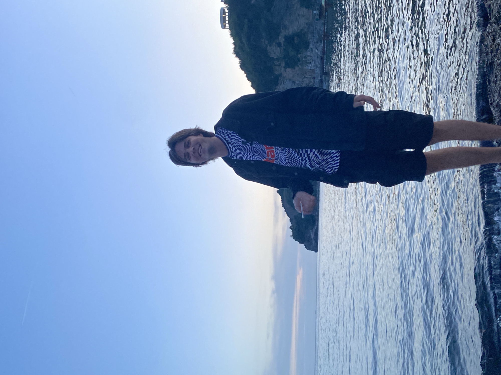 A medium shot depicts a fair-skinned male standing on a rocky shore in the city of Budva, Montenegro. He faces the camera, smiling slightly. He wears a black jacket, a shirt with a checkerboard pattern, and black shorts. He is positioned near the water's edge. The Adriatic Sea stretches out to the right, while a cliffside with greenery is in the background, also to the right. The sky, a pale blue, fills the left side of the frame.