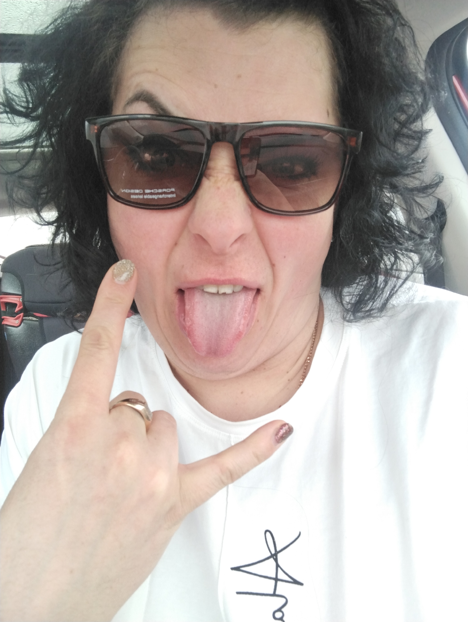 A close-up selfie captures an adult individual, likely female, with dark, curly hair and dark eyebrows, making a playful expression. The person is sticking out their tongue, scrunching their nose, and looking towards the camera while wearing dark brown, rectangular sunglasses. Through the left lens of the sunglasses, the reversed text "PORSCHE DESIGN" and "lens is polarized" is visible.
The subject is wearing a plain white t-shirt, with a stylized black drawing or logo partially visible at the bottom right. Both hands are raised, forming a "rock on" or "devil horns" gesture. The left hand, prominently in the foreground, has a gold-toned ring on the ring finger, and the index finger features glittery gold nail polish. The pinky finger of the right hand also shows a darker glittery nail polish. A thin, gold-colored chain necklace is visible around the neck.
The background suggests the photo was taken inside a car, possibly from the driver or passenger seat, with parts of a car seat and reflections on a window visible. The lighting is bright, indicating it is daytime. The overall tone of the image is humorous and informal. The location context is Izhevsk, Russia.
