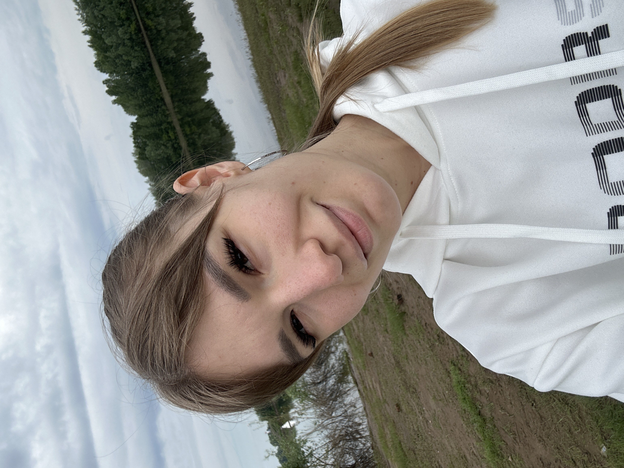 The image features a close-up, rotated portrait of a young woman, likely a selfie. Her head is tilted, and she looks towards the camera with a subtle, closed-mouth smile. She has long, light brown hair, some strands falling across her forehead and cheek. Her eyes are partially closed, accentuated by long, dark eyelashes. She wears a white hooded sweatshirt (hoodie), on which the brand name "UNDER ARMOUR" is visible in black and white stylized text, specifically "R ARMOUR" can be clearly seen. A small, silver hoop earring is visible in her left ear.
The scene is set outdoors, under a cloudy, overcast sky, suggesting daytime. In the background, a distinct line of tall, slender green trees, possibly poplars, runs vertically across the frame. The ground in the mid-to-foreground is a mix of sparse green grass and bare, earthy patches. The overall atmosphere is calm and natural. The location context indicates this scene is in Chaikovsky, Russia.
