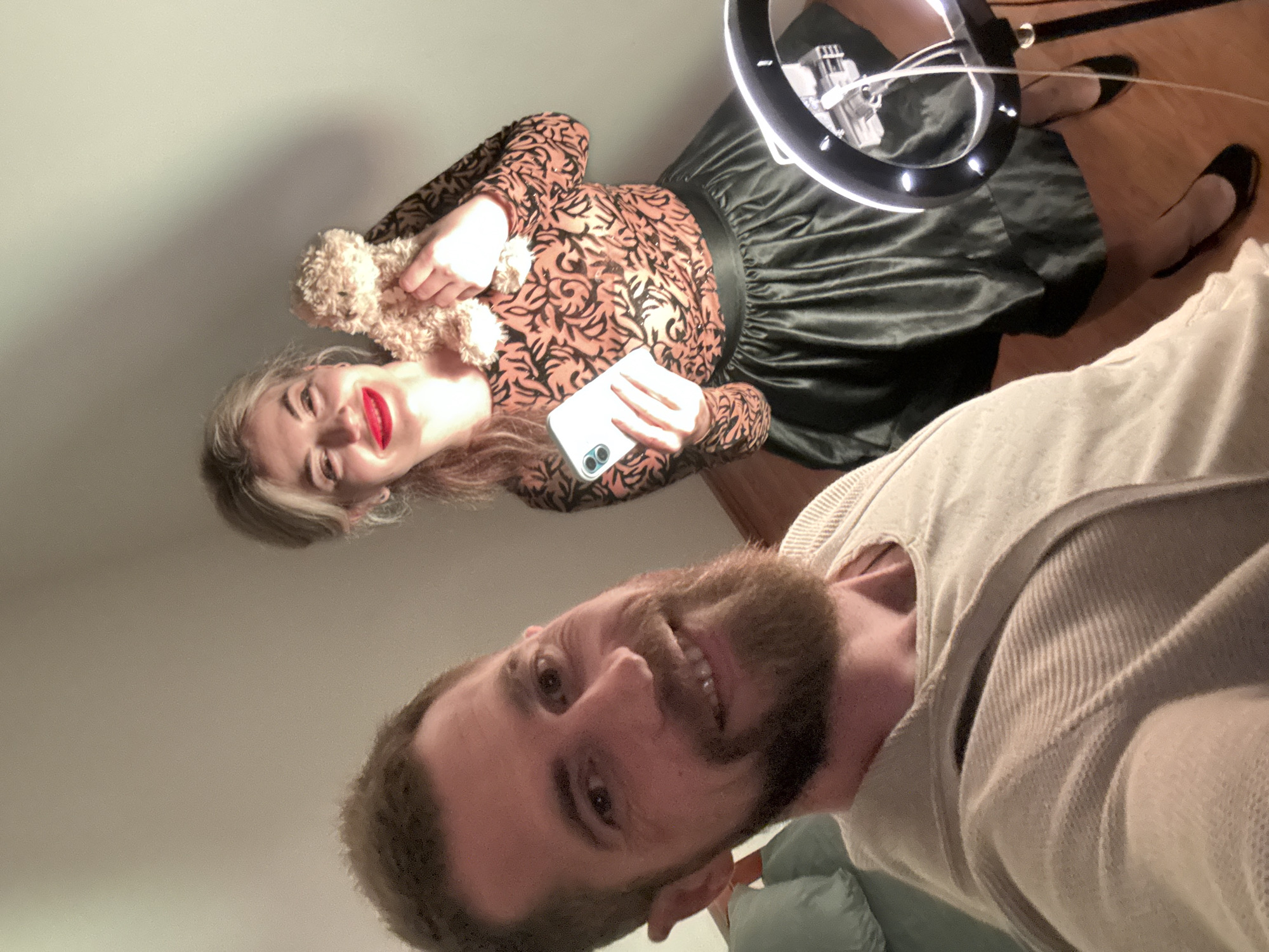 An indoor scene in Budva, Montenegro, depicts two individuals posing for a selfie or photograph. In the foreground, slightly to the left, an adult male with a brown beard and short brown hair smiles directly at the camera. He is wearing a light beige, ribbed long-sleeved top. Behind him, to the right, an adult female with blonde hair styled upwards and bright red lipstick smiles towards the camera. She is wearing a long-sleeved top with a dark patterned design on a reddish-orange background and a dark, possibly black, pleated midi-skirt. She holds a small, light-brown, fluffy stuffed animal in her left hand and a white smartphone in her right hand. A black ring light, illuminated, is positioned to the lower right of the frame, partially obscuring her feet which are clad in black flats on a wooden floor. The background consists of a plain light-colored wall and a glimpse of a light-green cushion on the far left. The precise location within Budva is not discernible beyond it being an indoor residential or commercial setting.