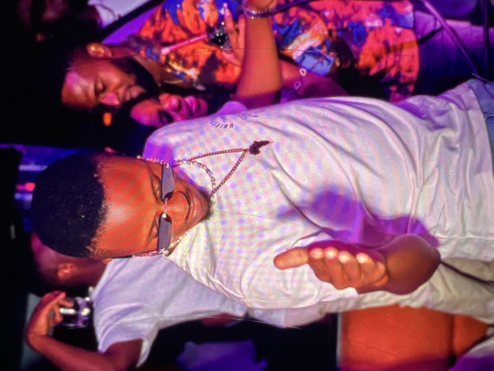 In Lagos, Nigeria, within a dimly lit establishment, likely a club or lounge, a man in the foreground wearing a white t-shirt and sunglasses is captured mid-gesture, with his right hand extended and thumb up. He is adorned with a gold chain featuring a pendant shaped like the continent of Africa. Behind him, another man in a vibrantly colored shirt and a third individual are visible, suggesting a social gathering. The presence of red seating and purple lighting indicates an indoor entertainment venue.