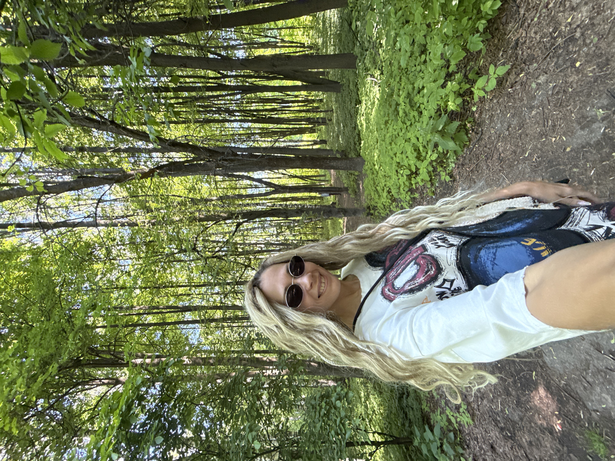 An adult female is depicted in a selfie taken outdoors in a sunlit forest or park in Rusanovo, Russia. The subject has long, wavy, light blonde hair and wears dark, oval-shaped sunglasses with a thin metallic frame. She is smiling, revealing her teeth. Her attire consists of a white t-shirt featuring a large graphic design with red lips, text, and blue/black elements. A black strap is visible over her right shoulder. She holds the camera with her right arm extended, angling her body slightly towards the lens. The background is a dense woodland area with numerous tall trees and lush green foliage, suggesting a summer environment. Bright sunlight filters through the tree canopy, creating dappled light on the forest floor. A dirt path, partially covered with fallen leaves and sparse vegetation, is visible in the foreground and extends into the background. To the right of the path, the ground is covered with a thick carpet of small, vibrant green leafy plants. A faint, distant figure in a light-colored top is discernible walking away on the path further into the woods.