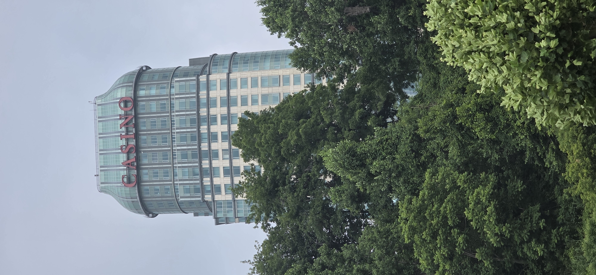 The image shows a tall, modern skyscraper with a distinctive curved, glass-enclosed top featuring the red word "CASINO" in large letters. The building has a segmented facade with numerous windows. It is partially obscured by lush green trees in the foreground, which fill the bottom half of the frame. The sky is overcast and appears a pale gray. The skyscraper is the Fallsview Casino Resort, located in Niagara Falls, Ontario, Canada. No people are visible in the image.
