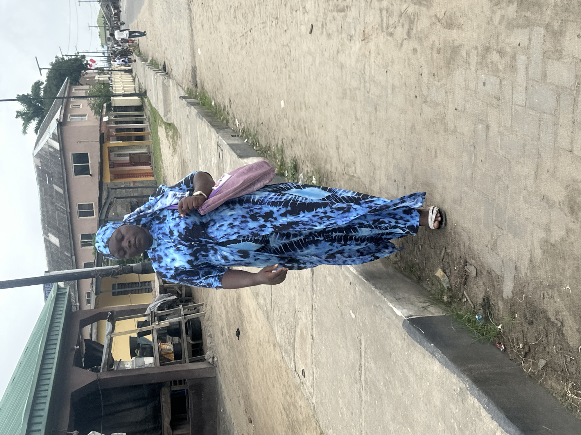 A full shot captures a woman walking towards the camera on a dusty street in Badagry, Nigeria. She is wearing a flowing blue and black patterned dress with a matching headscarf. A light purple bag hangs from her right shoulder. The street is lined with concrete sidewalks and some overgrown vegetation. In the background, buildings with metal roofs and weathered facades suggest a modest urban environment. A few other individuals are visible walking further down the street. The sky is overcast, contributing to the muted lighting of the scene.