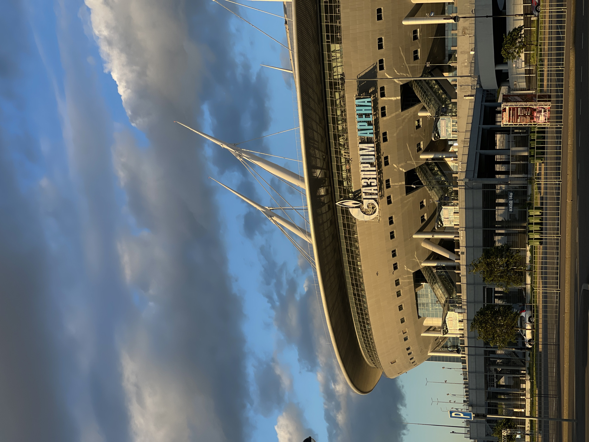 A low-angle exterior shot captures the Gazprom Arena stadium under a dramatic sky. The stadium's distinctive curved facade is illuminated by warm, late-afternoon sunlight, casting long shadows. The architectural features include a complex roof structure supported by angled white pylons. The name "ГАЗПРОМ АРЕНА" is prominently displayed in large, illuminated lettering across the stadium's upper section. Below the main structure, a multi-level concourse area with visible escalators and staircases is partially obscured by supporting columns.
In the foreground, a street runs parallel to the stadium, separated by a metal fence. A silver car is parked on the left side of the street, and two trees stand behind the fence. The sky is a dynamic mix of dark, heavy clouds and patches of bright blue, with a few white clouds catching the light. A blue parking sign is visible on the left. The overall scene suggests a large, modern sports venue in an urban setting.