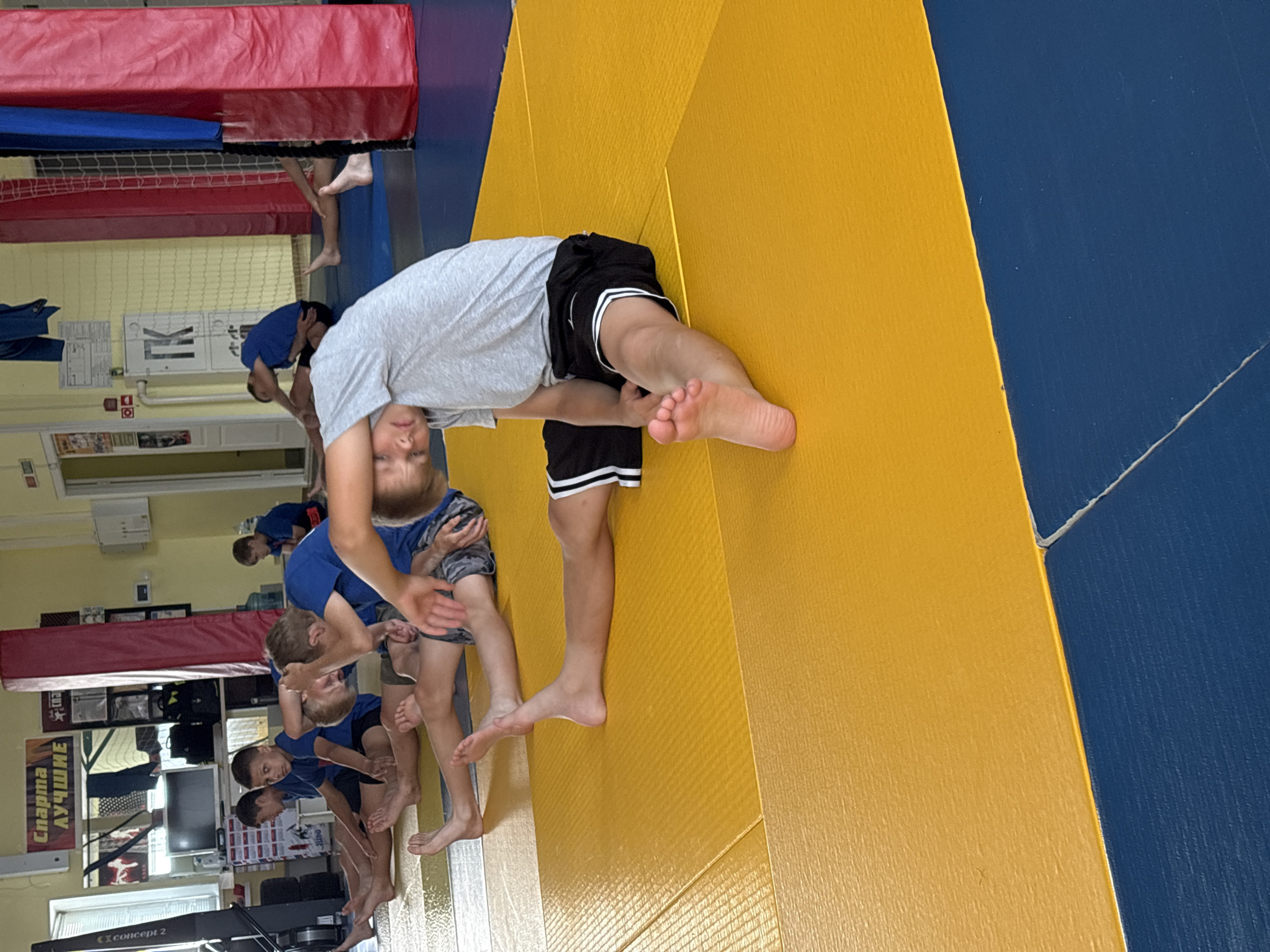 The image shows a group of young boys in athletic attire, practicing stretching exercises on a yellow and blue mat in what appears to be a gymnasium. The boys are in various seated stretching positions, with one boy in the foreground prominently displaying a seated forward bend. In the background, there are posters on the wall, a mirror, some gym equipment, and a large red and blue padded structure. The environment suggests a sports training facility. The poster in the background reads "Спарта Лучшие" which translates to "Sparta The Best", indicating a connection to a sports club or school. Although not explicitly stated in the image, the text on the poster is in Russian and relates to sports clubs, and the gymnasium setting are consistent with a facility in Chaikovsky, Russia, which is known for its sports complexes, particularly for combat sports.