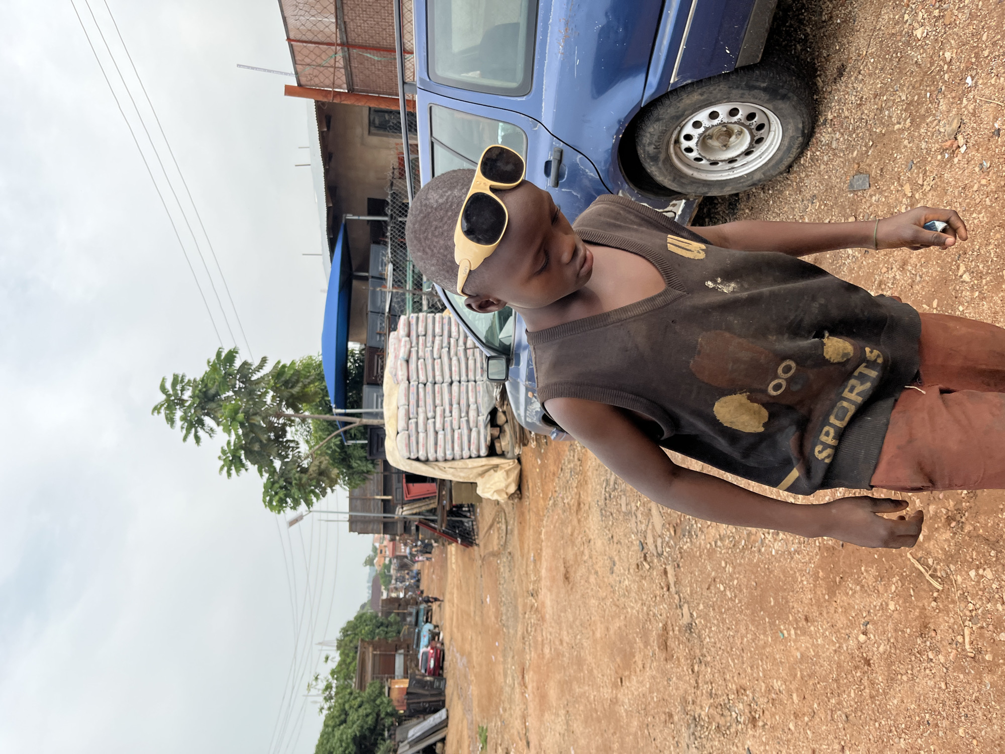 A young boy, wearing a black sleeveless shirt with a gold graphic and yellow-rimmed sunglasses, stands in a dusty outdoor area. He is holding a small, rectangular object in his right hand. To his left, a stack of white bagged goods, possibly cement, is visible. Behind the boy and the bags, a blue vehicle is parked. The ground is a mix of dirt and small stones. In the background, a row of buildings and trees lines a dirt road, with more vehicles and activity suggesting a market or busy thoroughfare. The sky is overcast, indicating a cloudy day. The overall scene depicts a bustling, somewhat rugged environment. The text "SPORTS" is visible on the boy's shirt.
